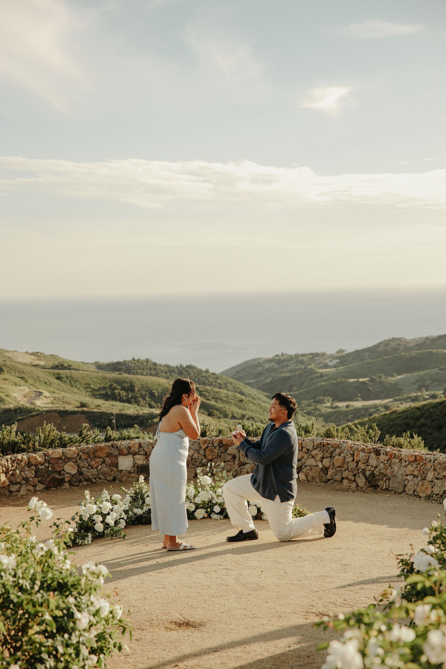 Man proposing on one knee to woman in white dress at scenic coastal overlook with ocean views and floral arrangements.