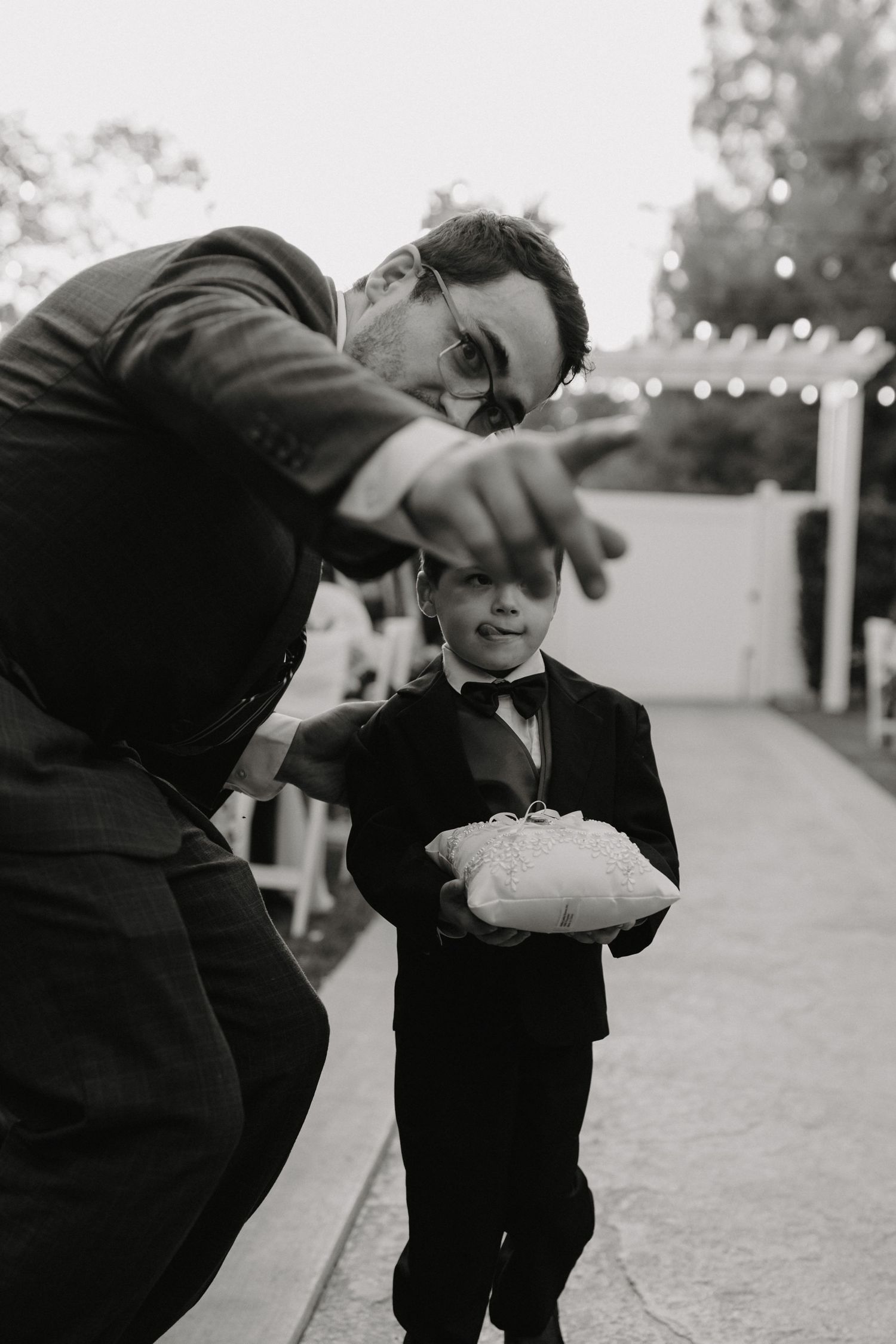 Black and white photo showing a ring bearer in a tuxedo holding a pillow while someone gestures during a wedding ceremony.