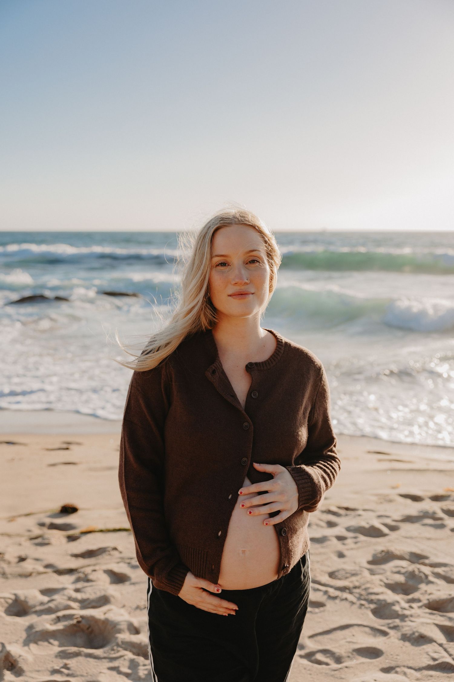 Expectant woman in brown shirt stands on sunny beach with ocean waves crashing behind her.