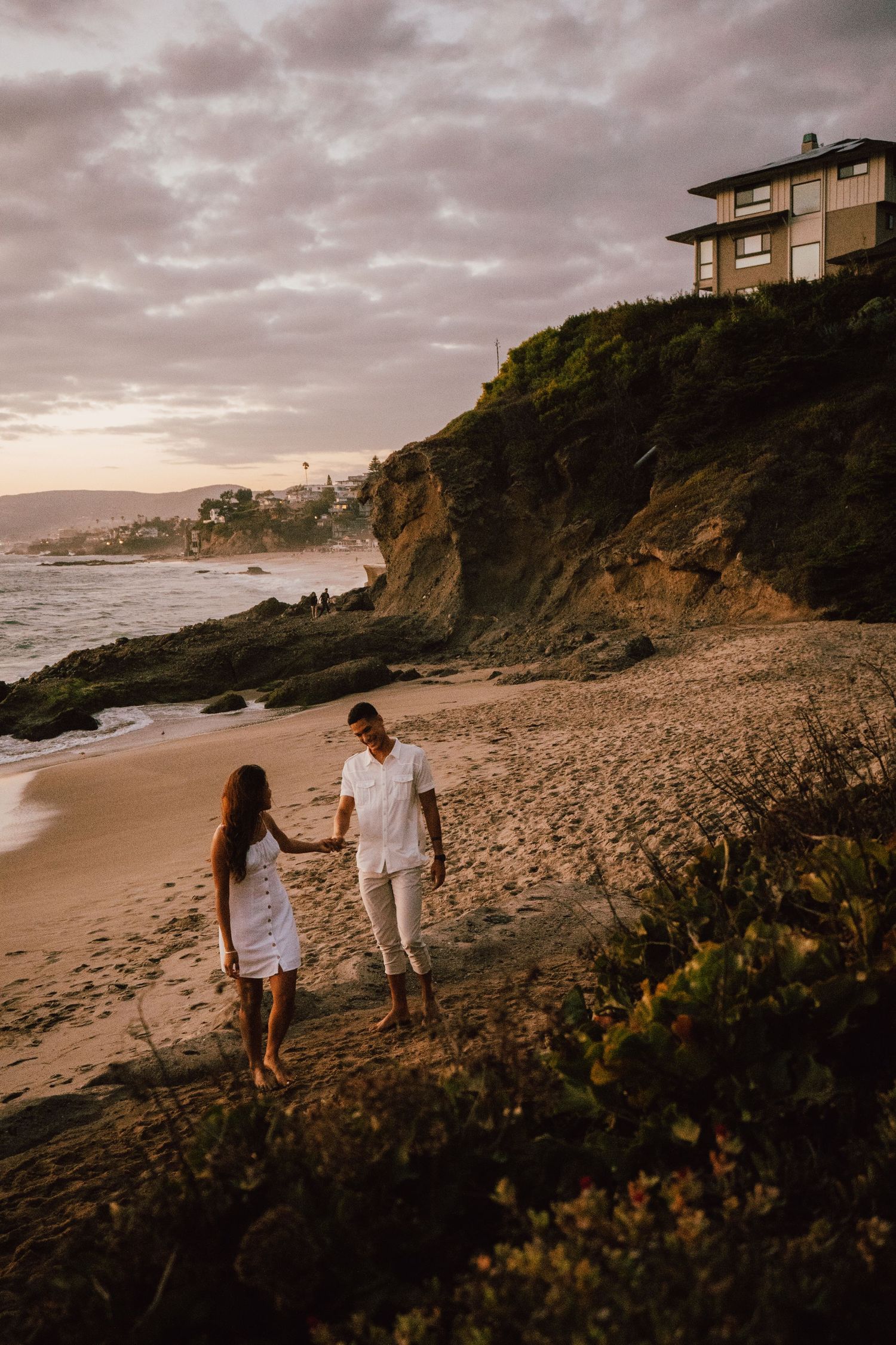 Couple walking on beach at sunset with cliffside homes in background.
