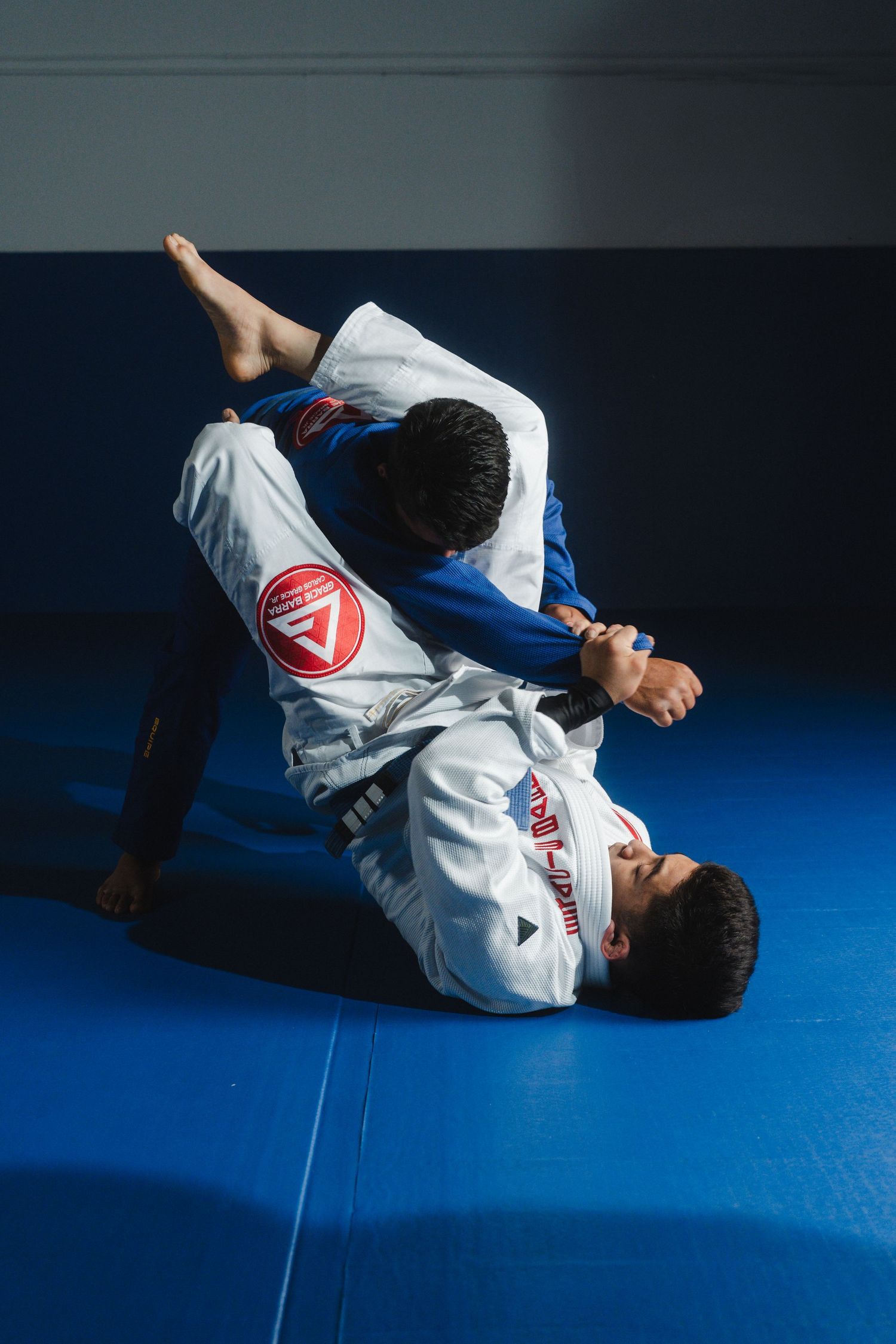 Two martial artists practicing Brazilian Jiu-Jitsu grappling techniques on blue mats.