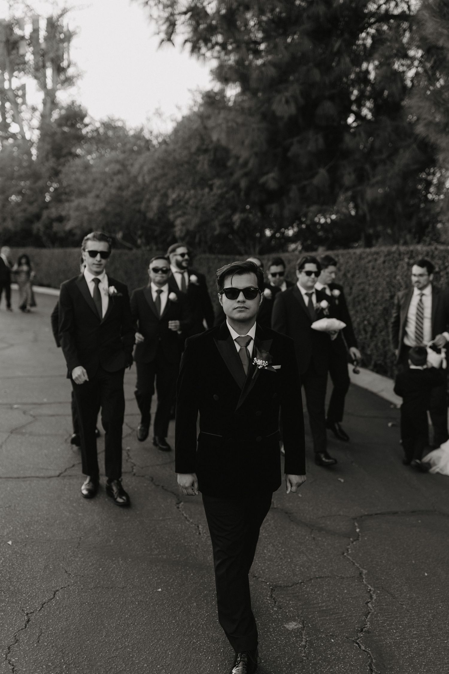 A group of people wearing dark suits and sunglasses walk together in a line down a path in black and white photography.