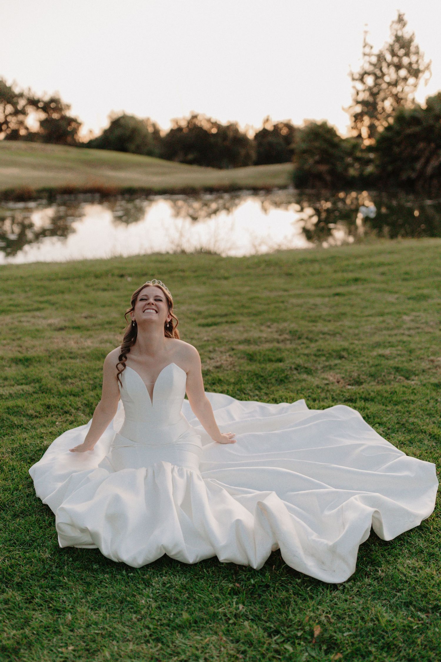 A bride in a flowing white wedding dress lies gracefully on green grass during golden hour sunset.