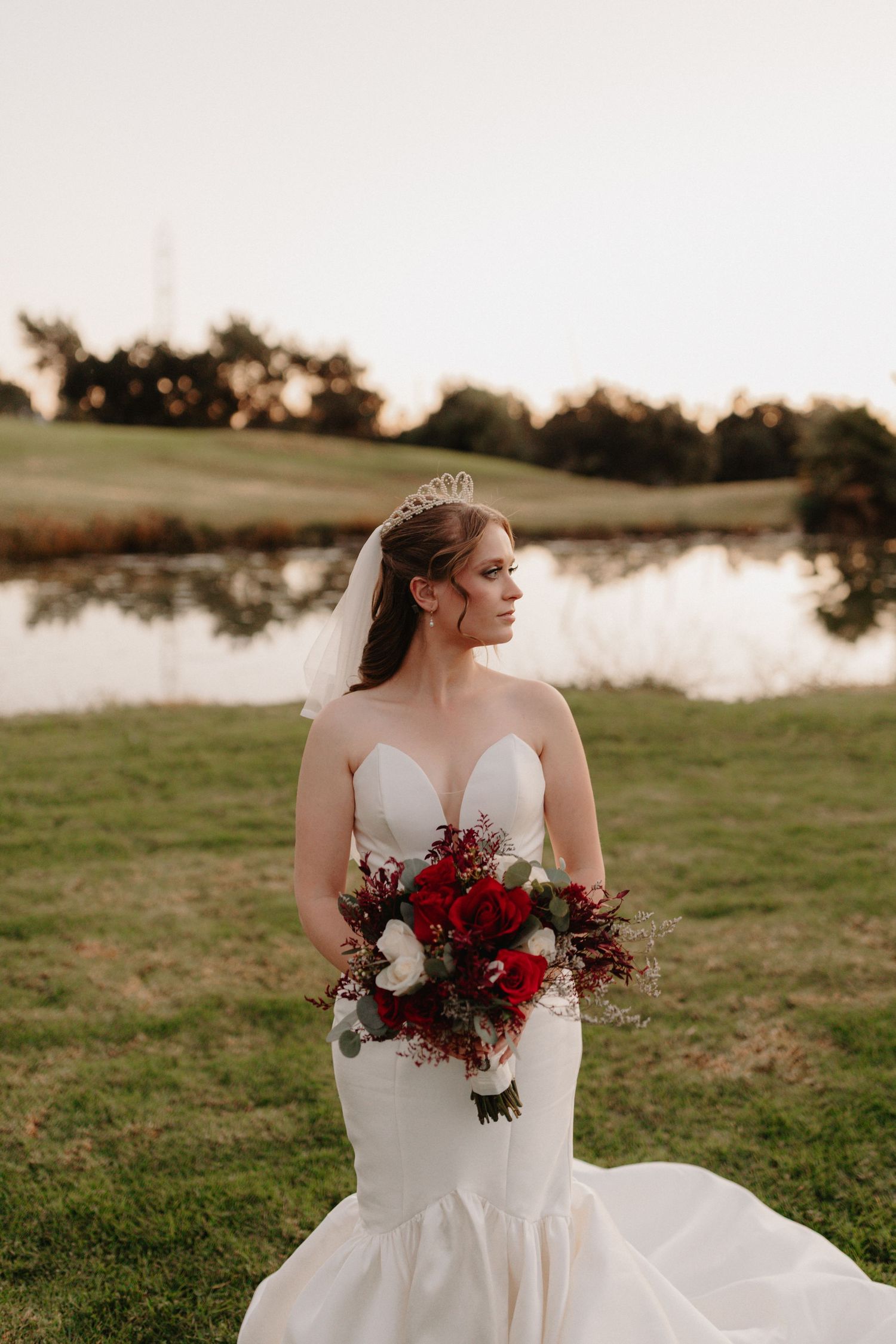 A bride in a white wedding dress holds a burgundy and white bouquet while standing near a lake at sunset.