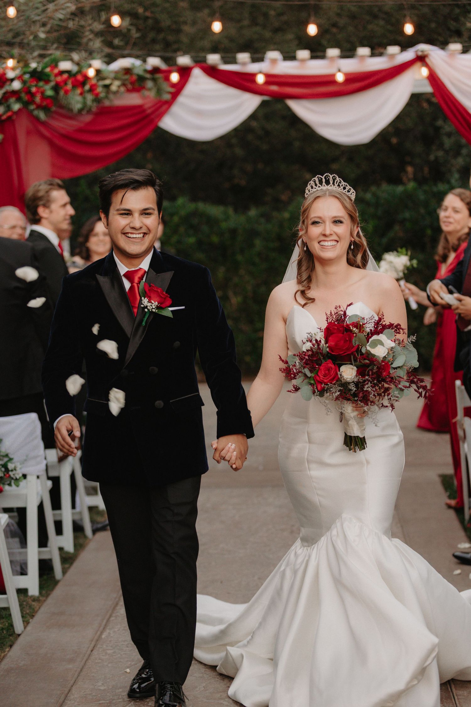 Wedding couple walks down aisle while guests throw white rose petals during outdoor ceremony with red decor.