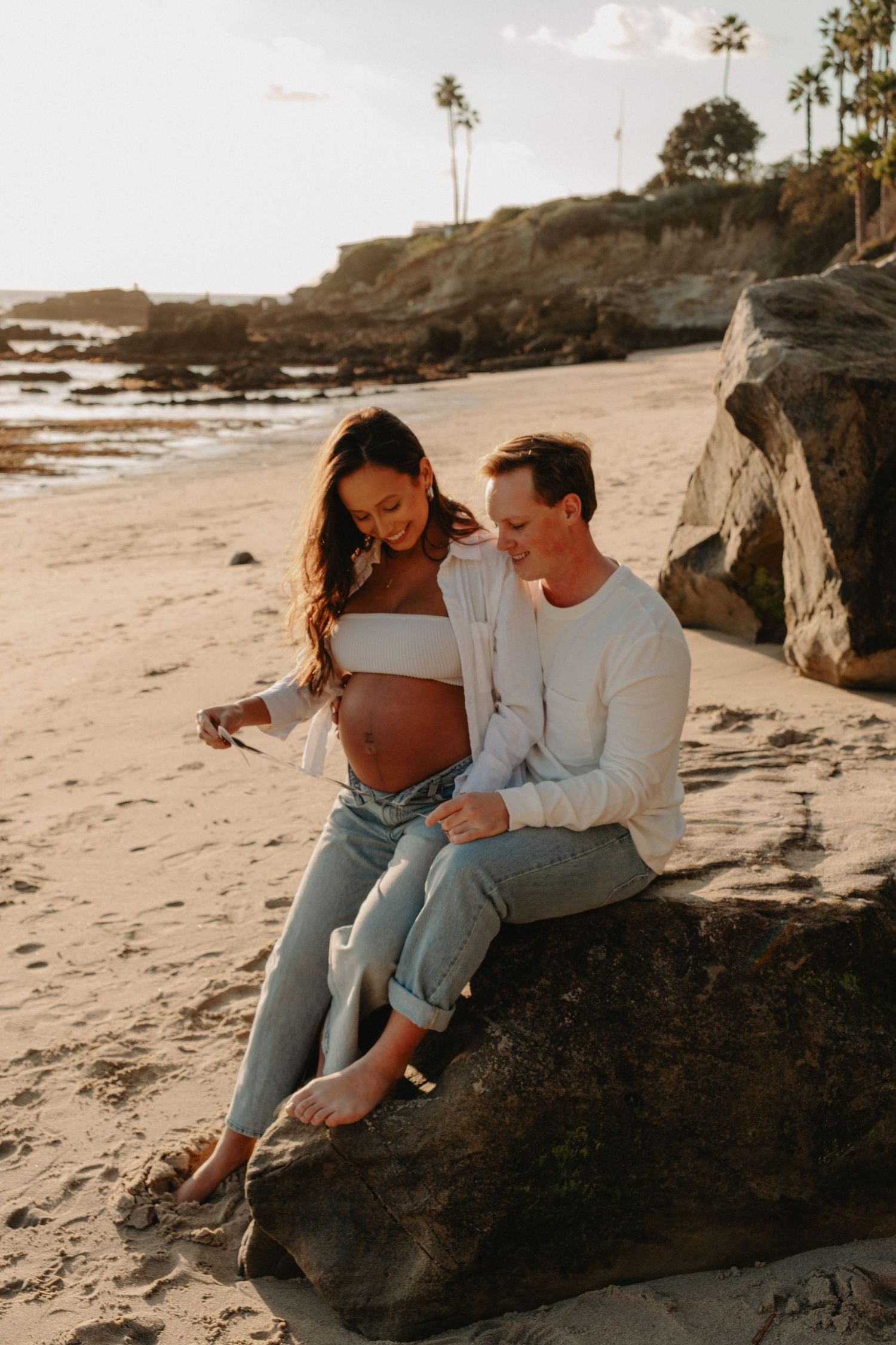 A couple sits together on coastal rocks at sunset sharing intimate moments on a sandy beach.