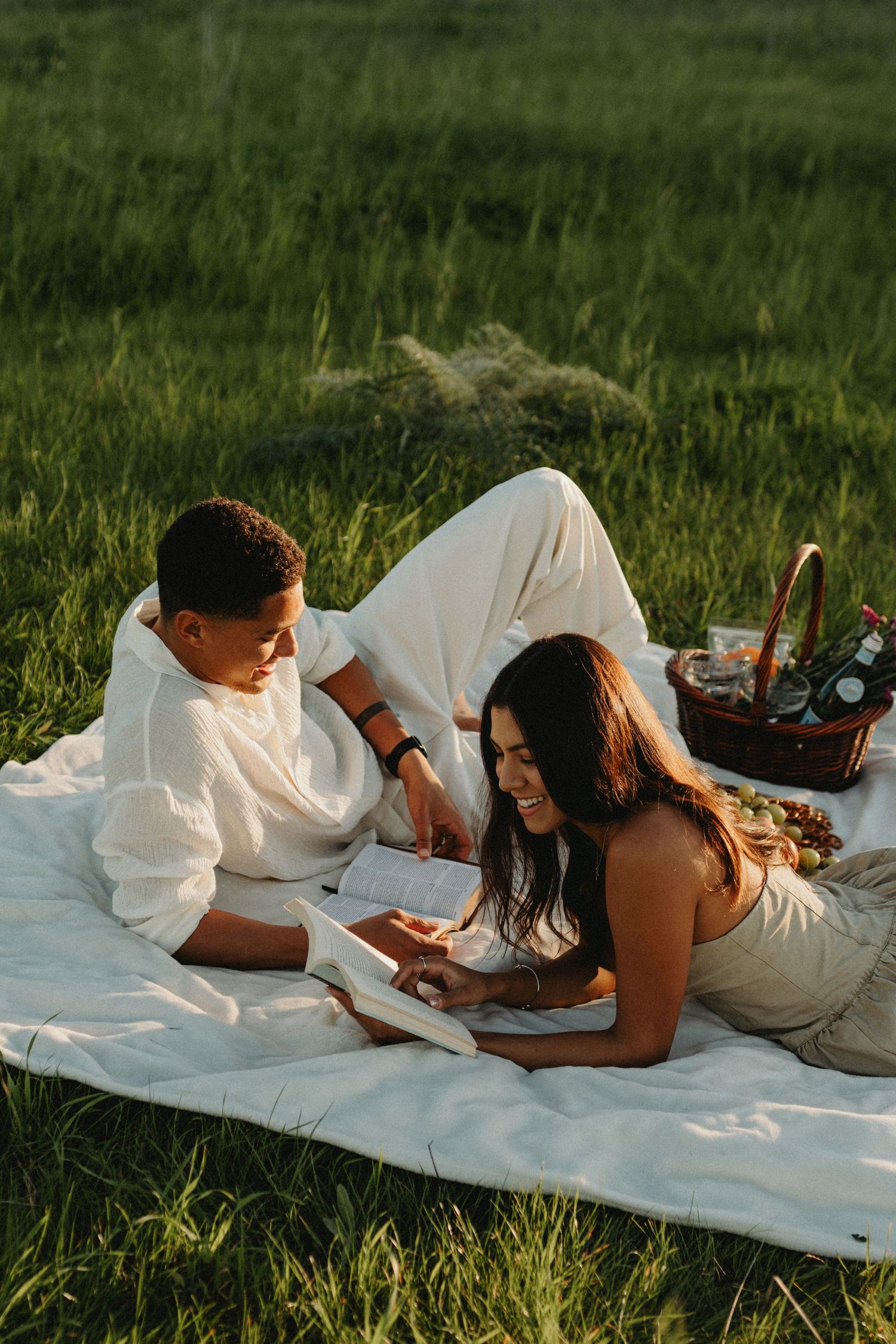 A couple lounges on a white blanket in a grassy field while reading books together on a sunny day.