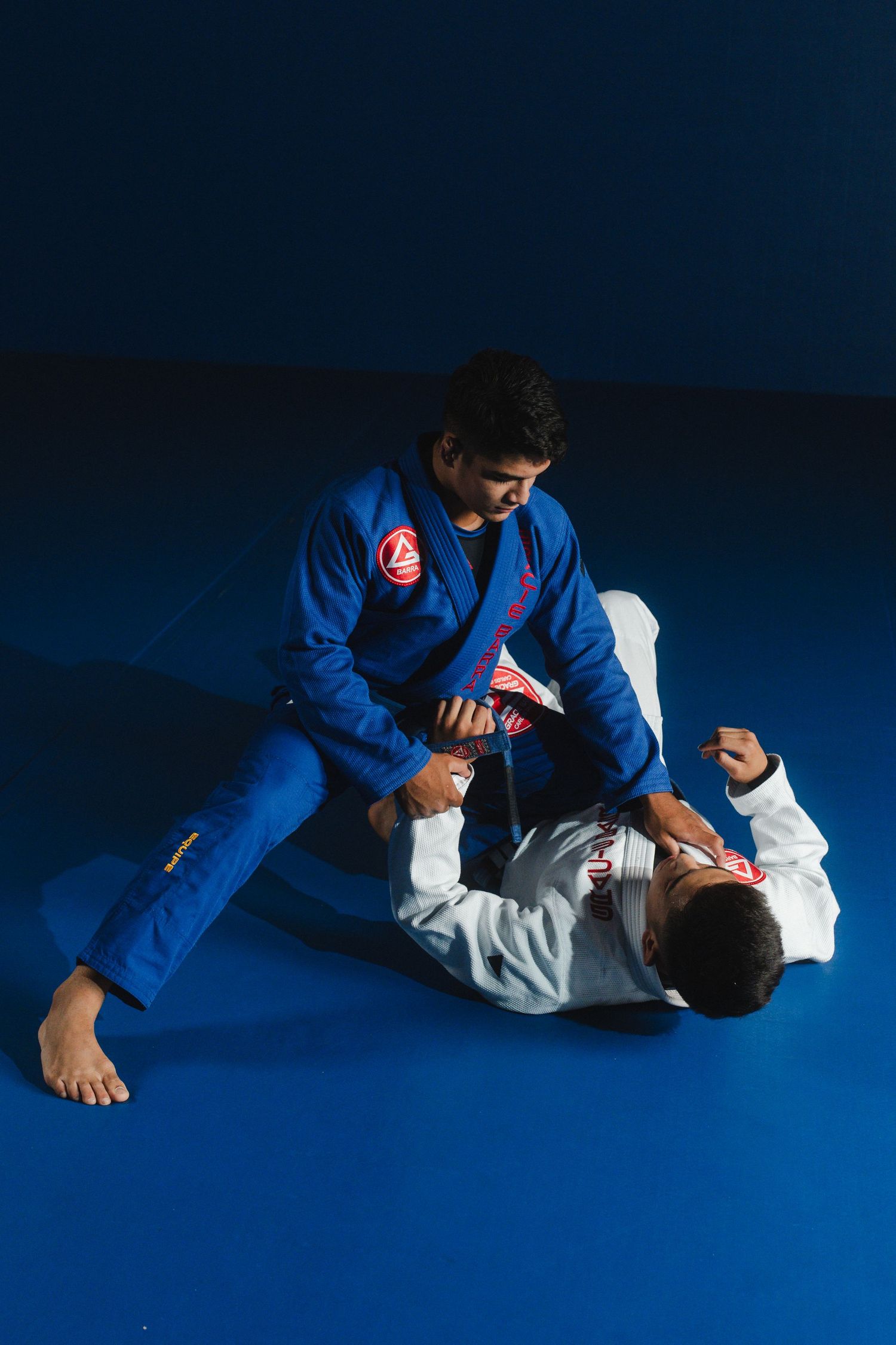 Two practitioners sparring in Brazilian Jiu-Jitsu with one wearing blue gi and other in white gi.