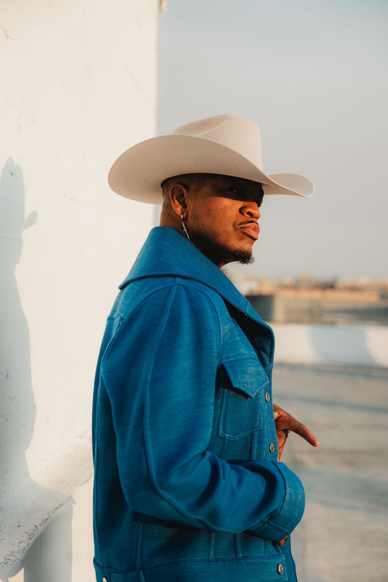 A person wearing a cowboy hat and blue denim jacket poses against a white wall.