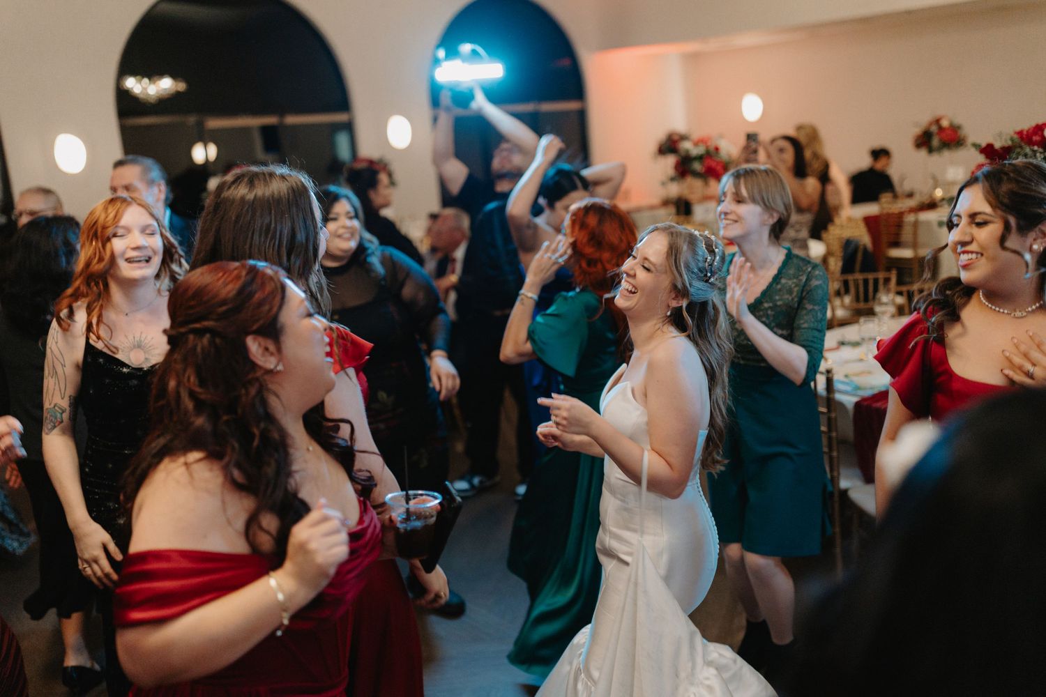 Wedding guests and a bride in formal attire dancing and celebrating at an indoor reception venue.