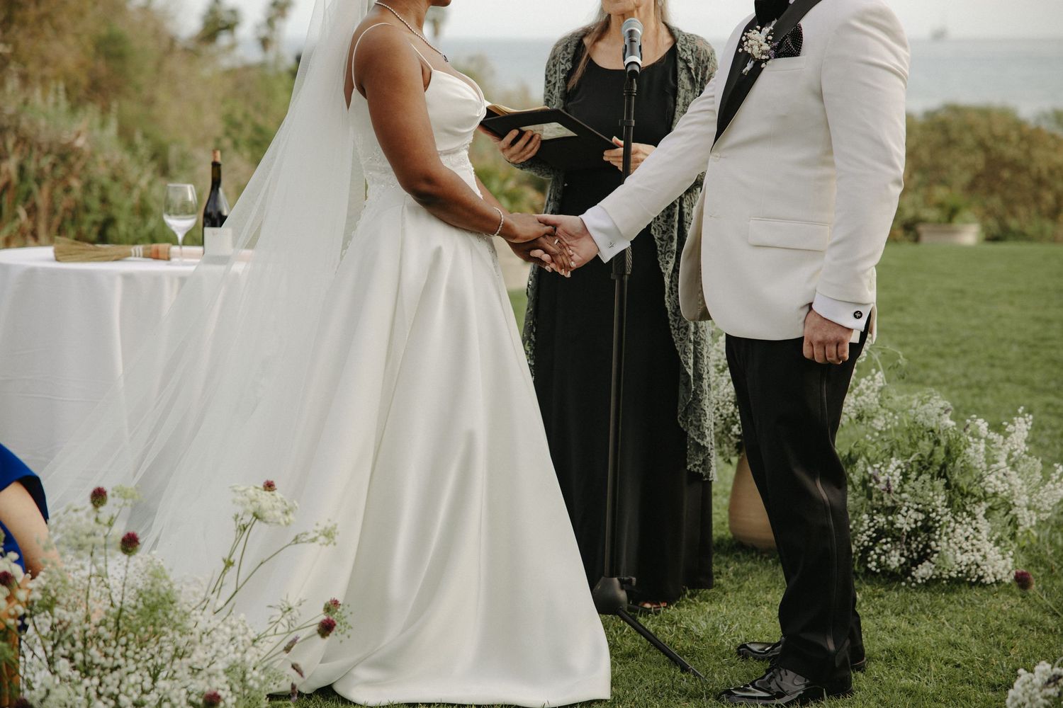 A couple holds hands during an outdoor wedding ceremony with a scenic countryside backdrop.