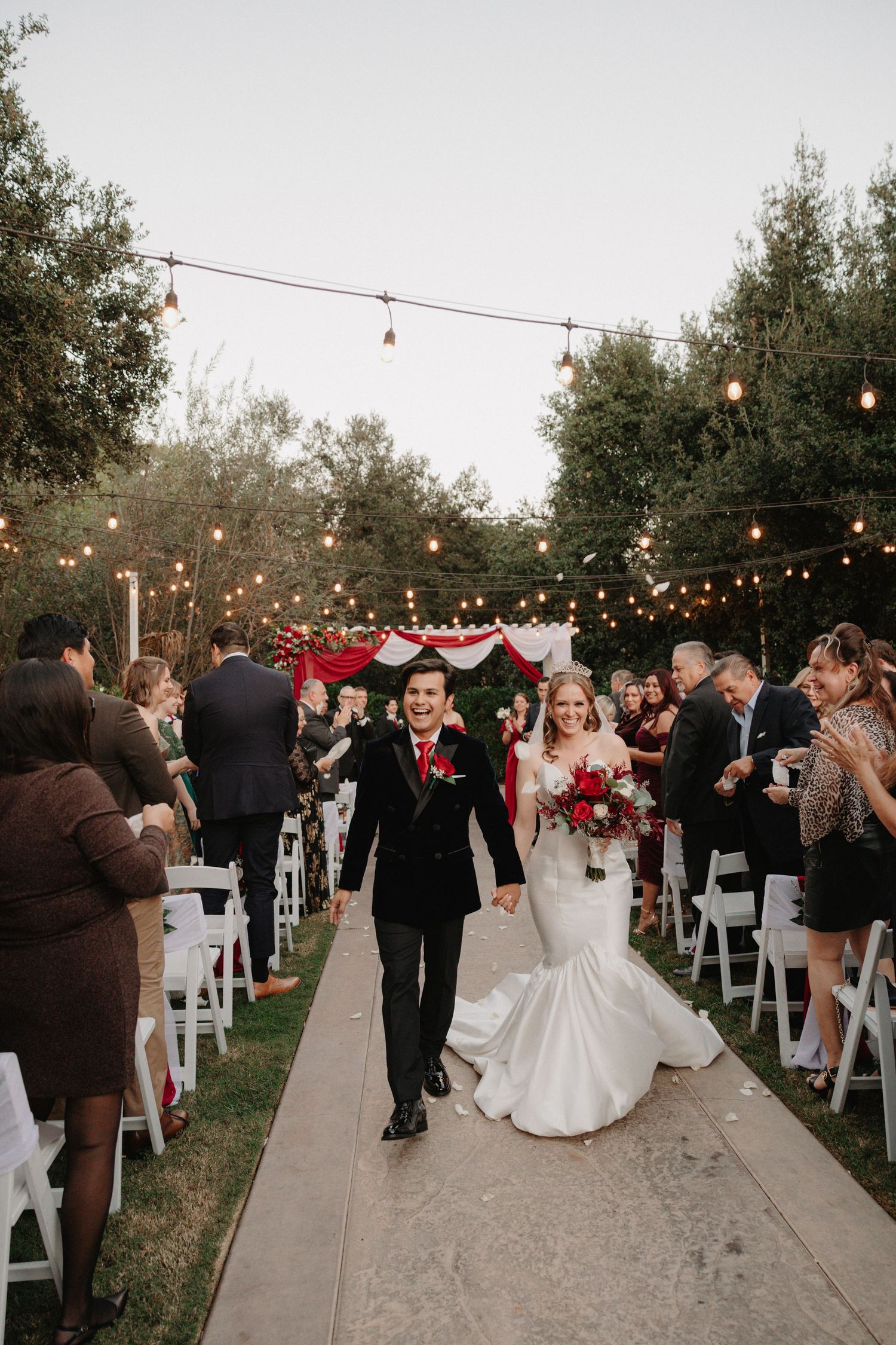Newlyweds walk down outdoor aisle under string lights while guests celebrate at garden wedding ceremony.