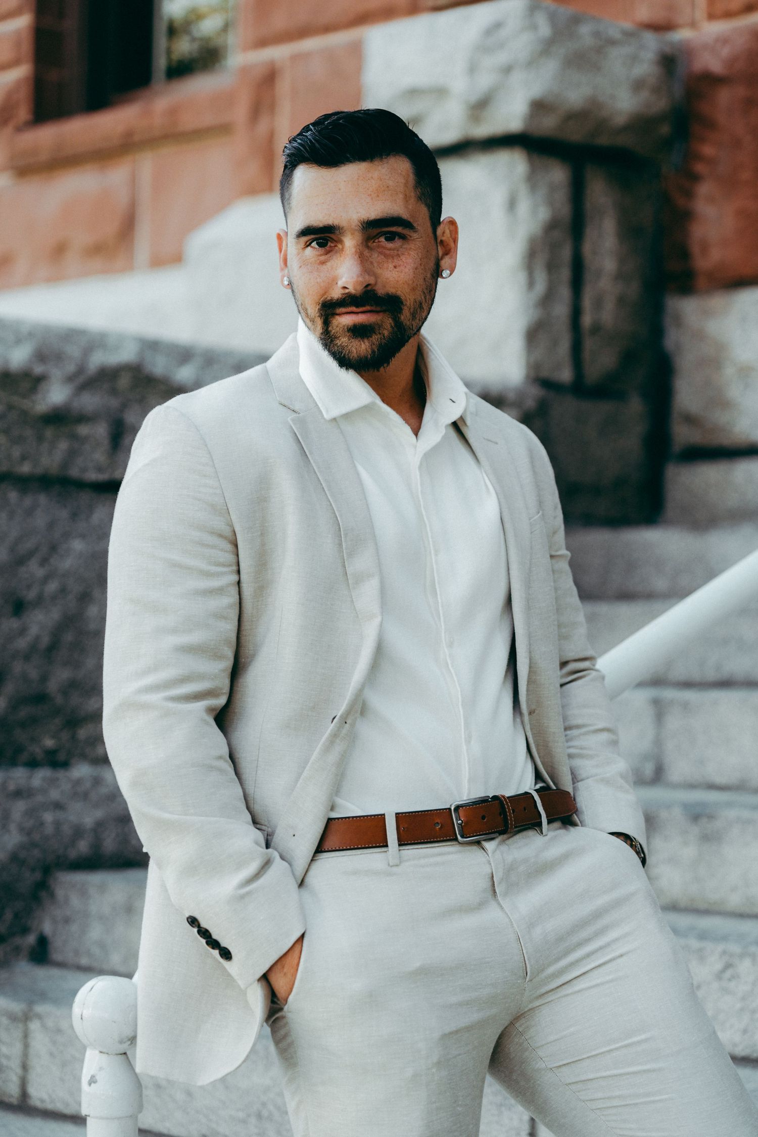 A groom in an all-white suit poses on stone steps near a brick building.