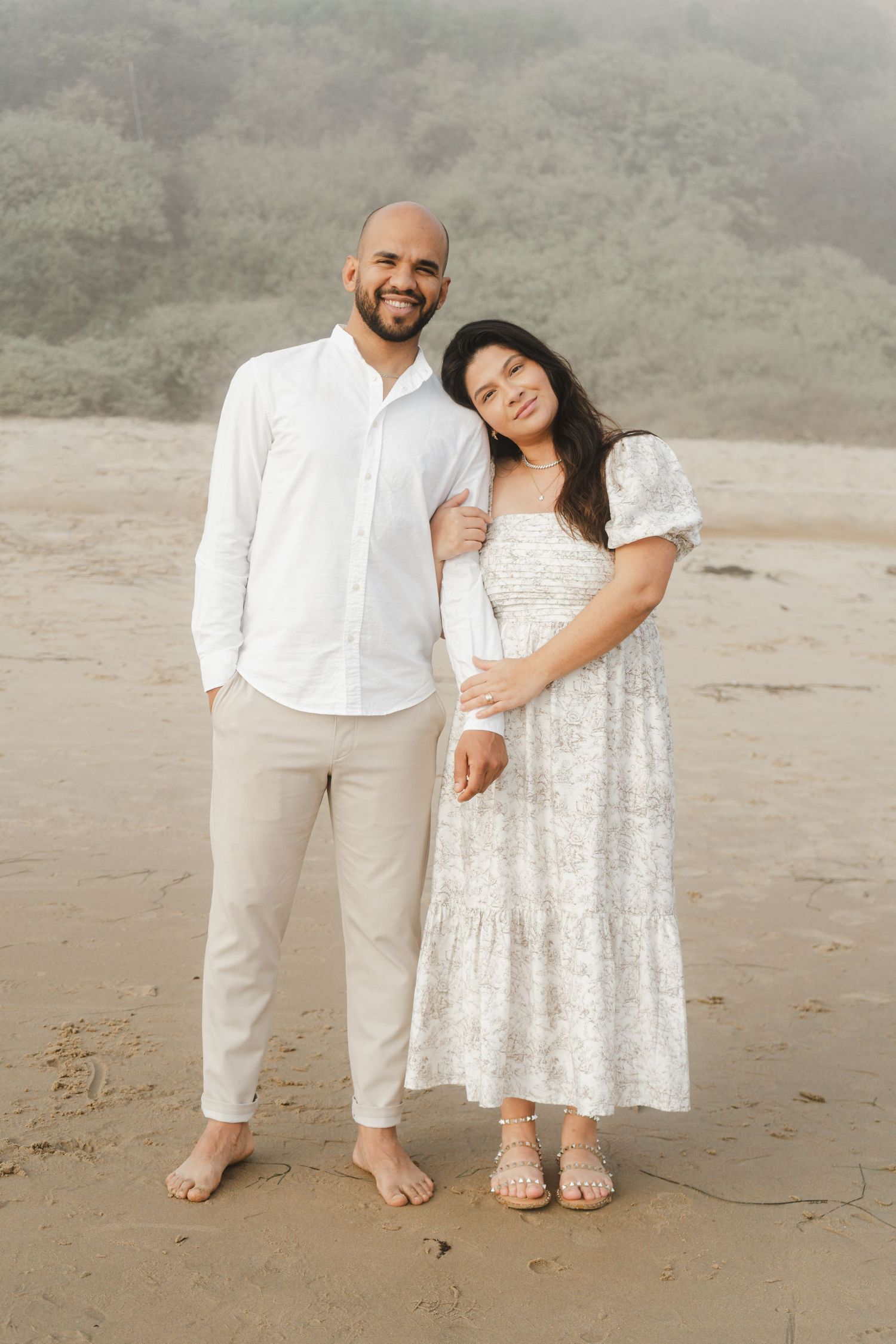 A couple in white outfits shares intimate moments on a misty beach during a romantic photoshoot.