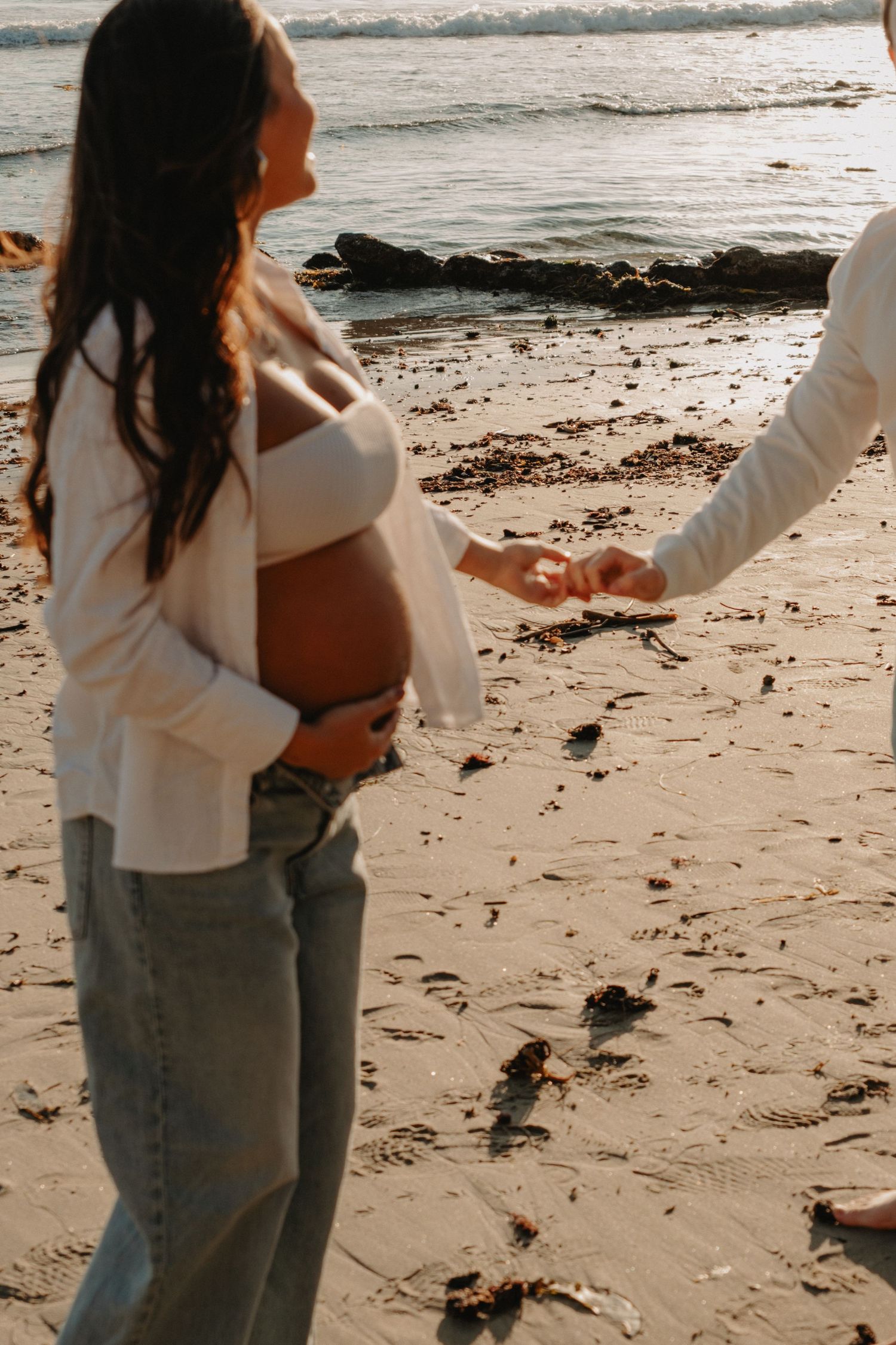 A couple embraces on a sandy beach at sunset with ocean waves in the background.