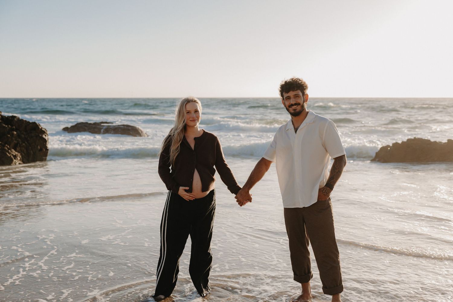 Couple walking hand in hand along a scenic beach at sunset with waves crashing on the shore.