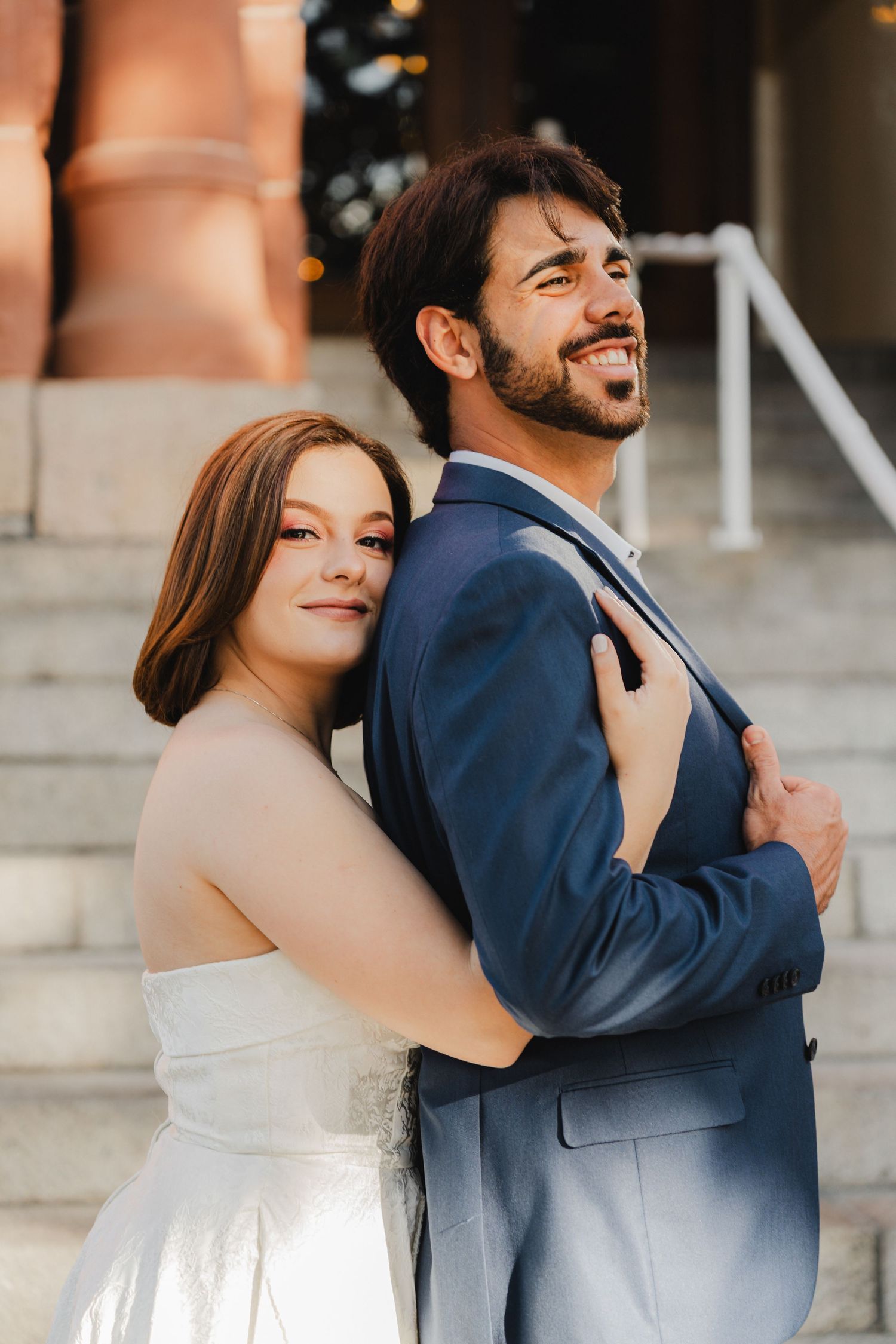 A couple embraces and laughs together on stone steps, with one wearing a navy suit and the other in white.