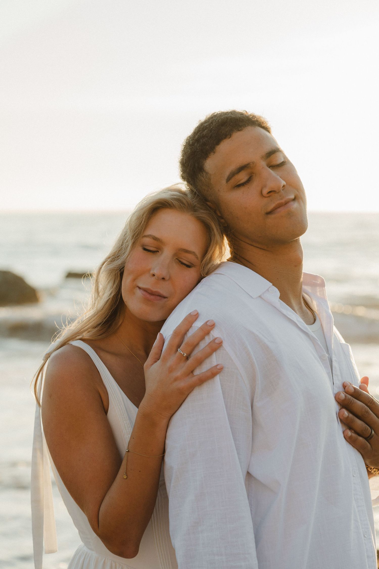 Romantic couple embraces on beach at sunset in white clothing with soft golden light.