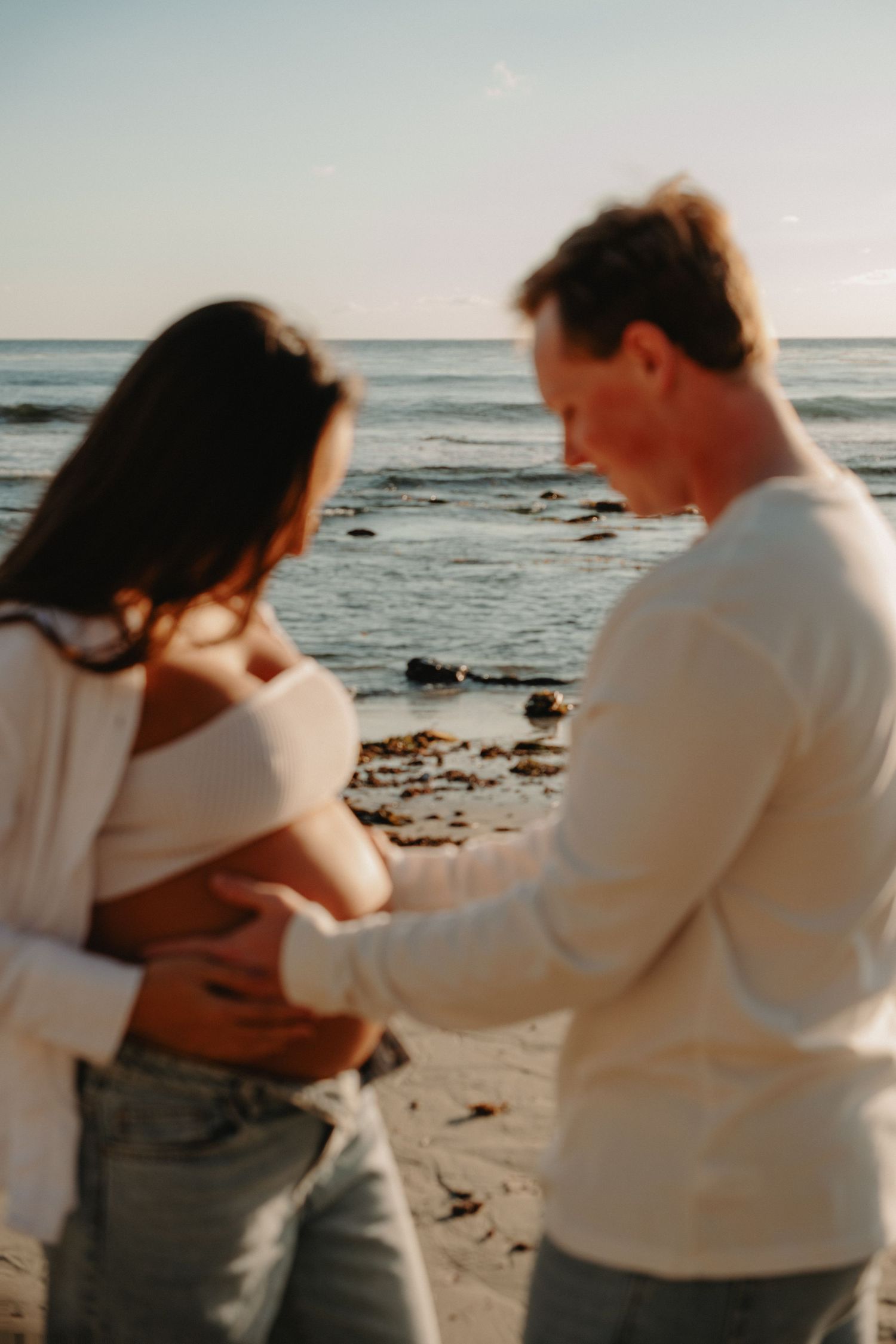 A couple in white sweaters share an intimate moment together on a rocky beach at sunset.