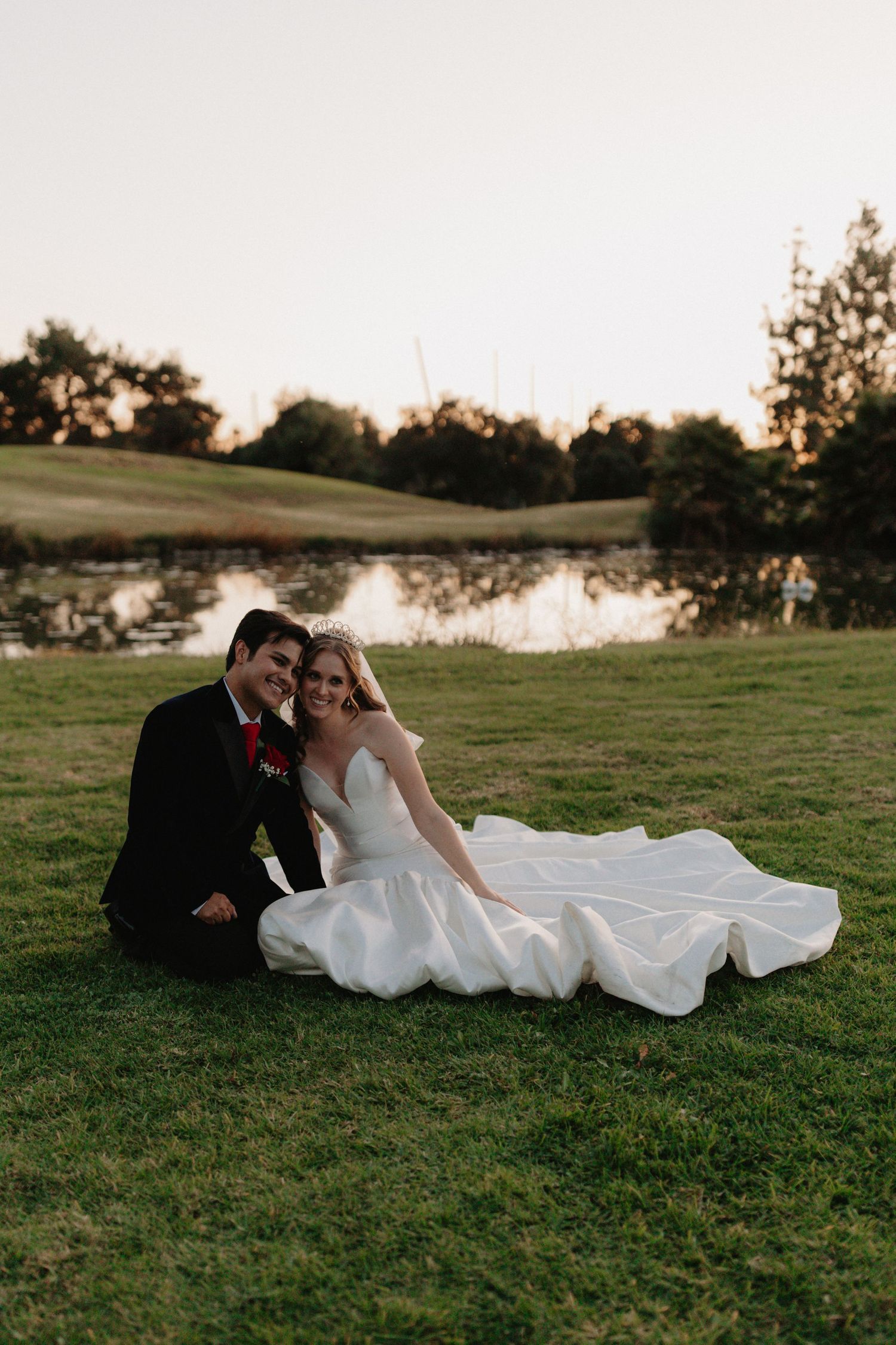 A couple in wedding attire lies together on a grassy hillside at sunset during their outdoor wedding photoshoot.