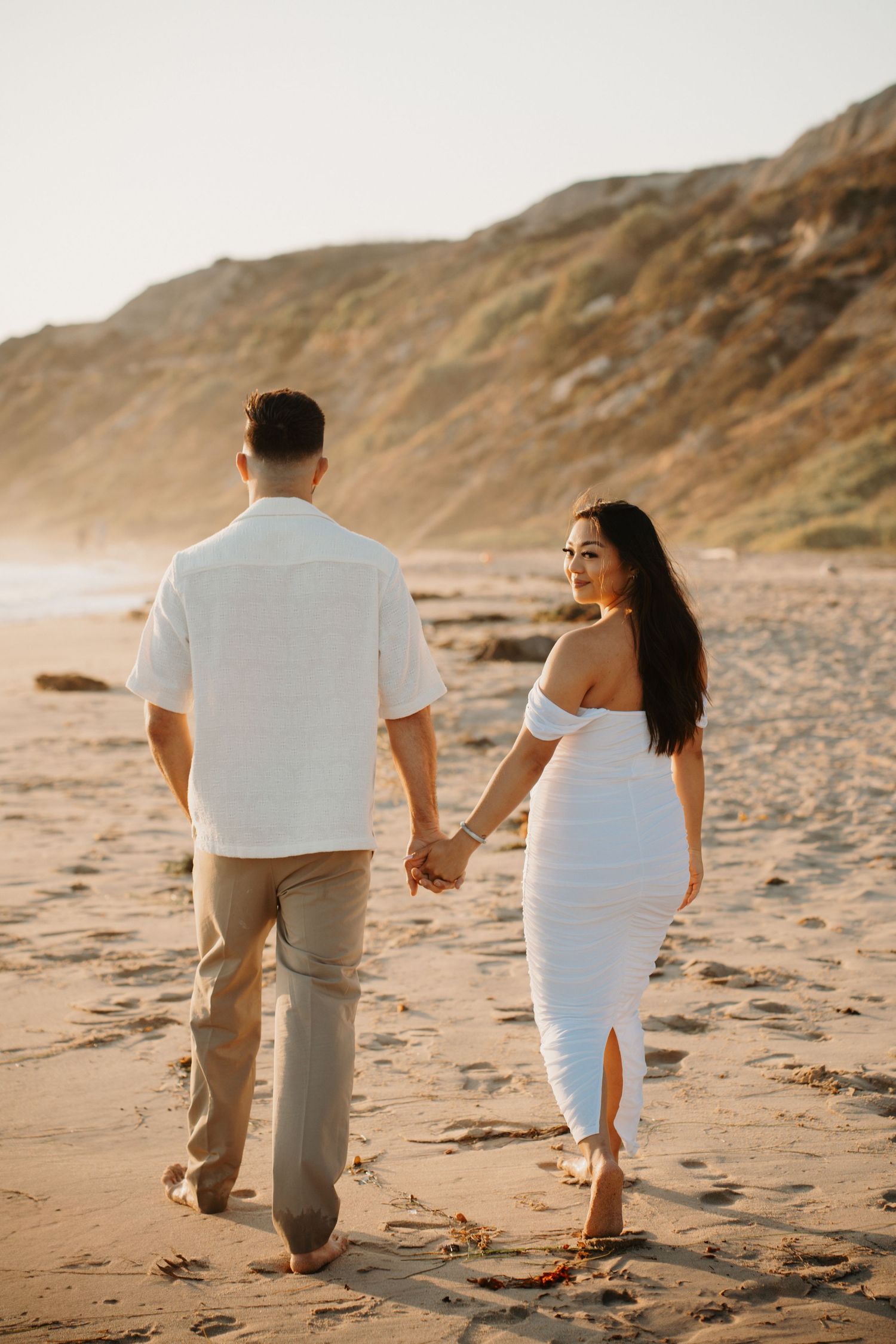 A couple walks hand in hand along a scenic coastal beach at sunset.