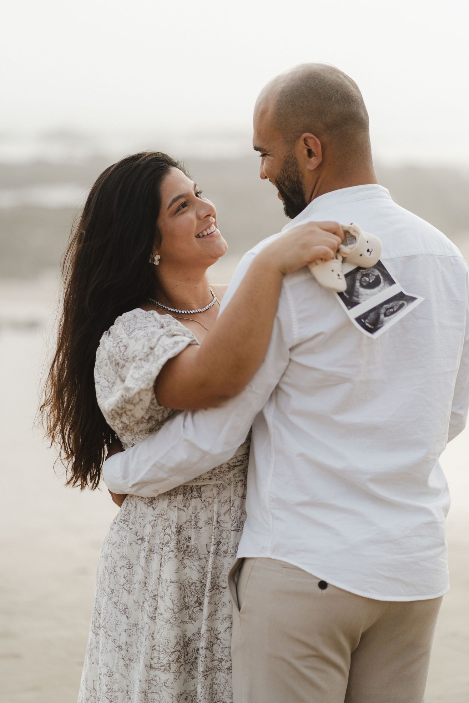 A couple shares a joyful embrace on a misty beach, wearing white and beige outfits.