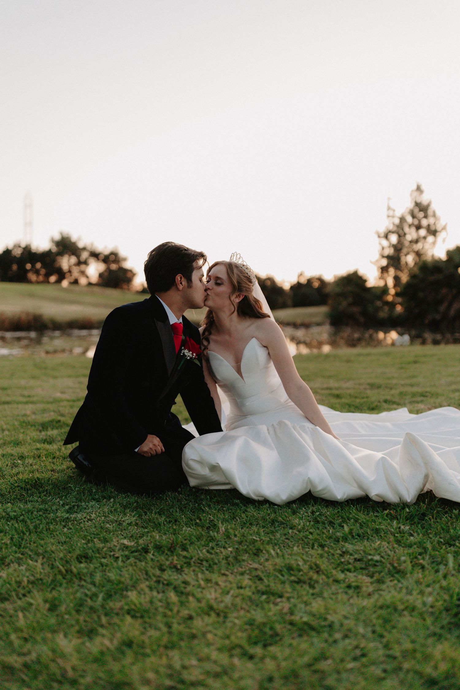 Newlywed couple shares romantic moment sitting on grass at sunset with bride's white dress flowing around them.