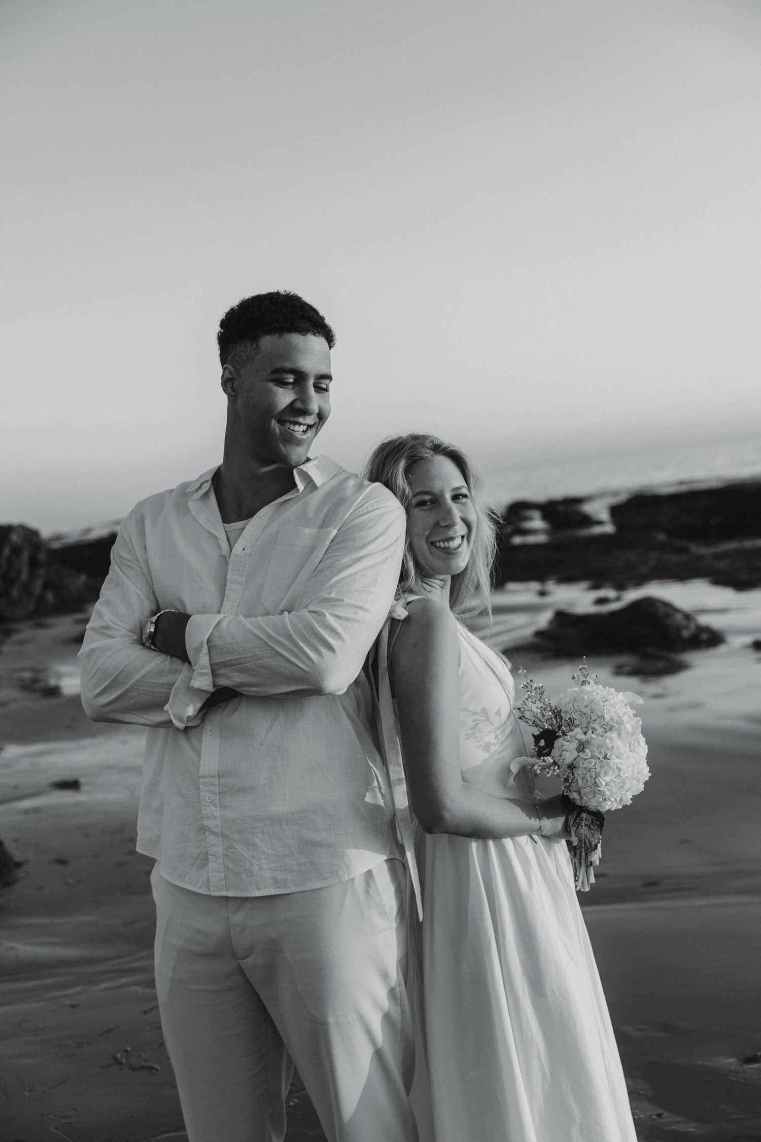 A romantic black and white beach photoshoot showing a couple in white attire embracing on a rocky coastline.