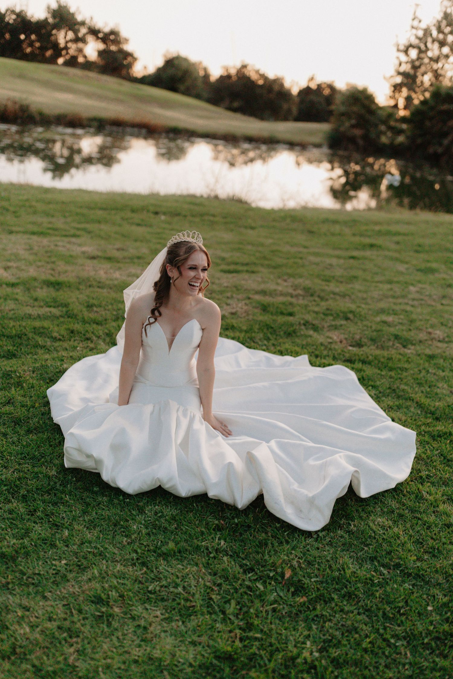A bride in a flowing white wedding dress reclines gracefully on lush green grass during a sunset photoshoot.
