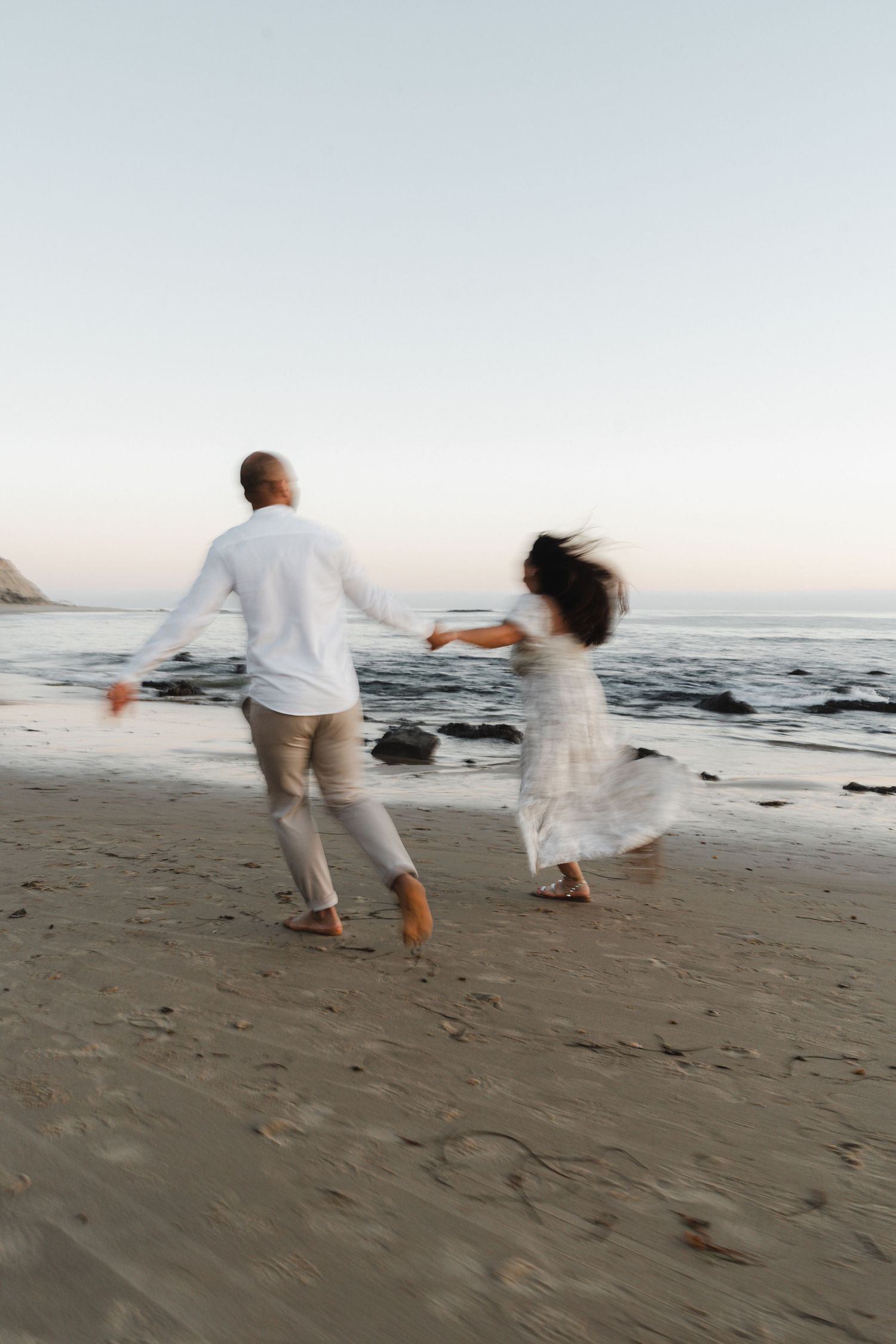 Two people in white outfits run playfully along a sandy beach at sunset with ocean waves in the background.