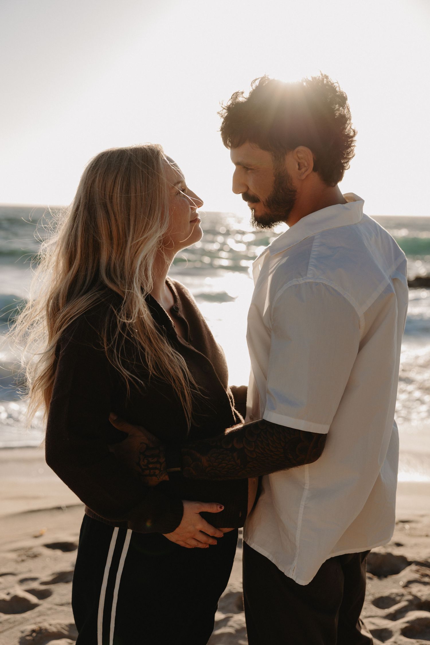 A romantic couple embraces on a sunny beach during golden hour as their silhouettes are backlit by the warm sunlight.