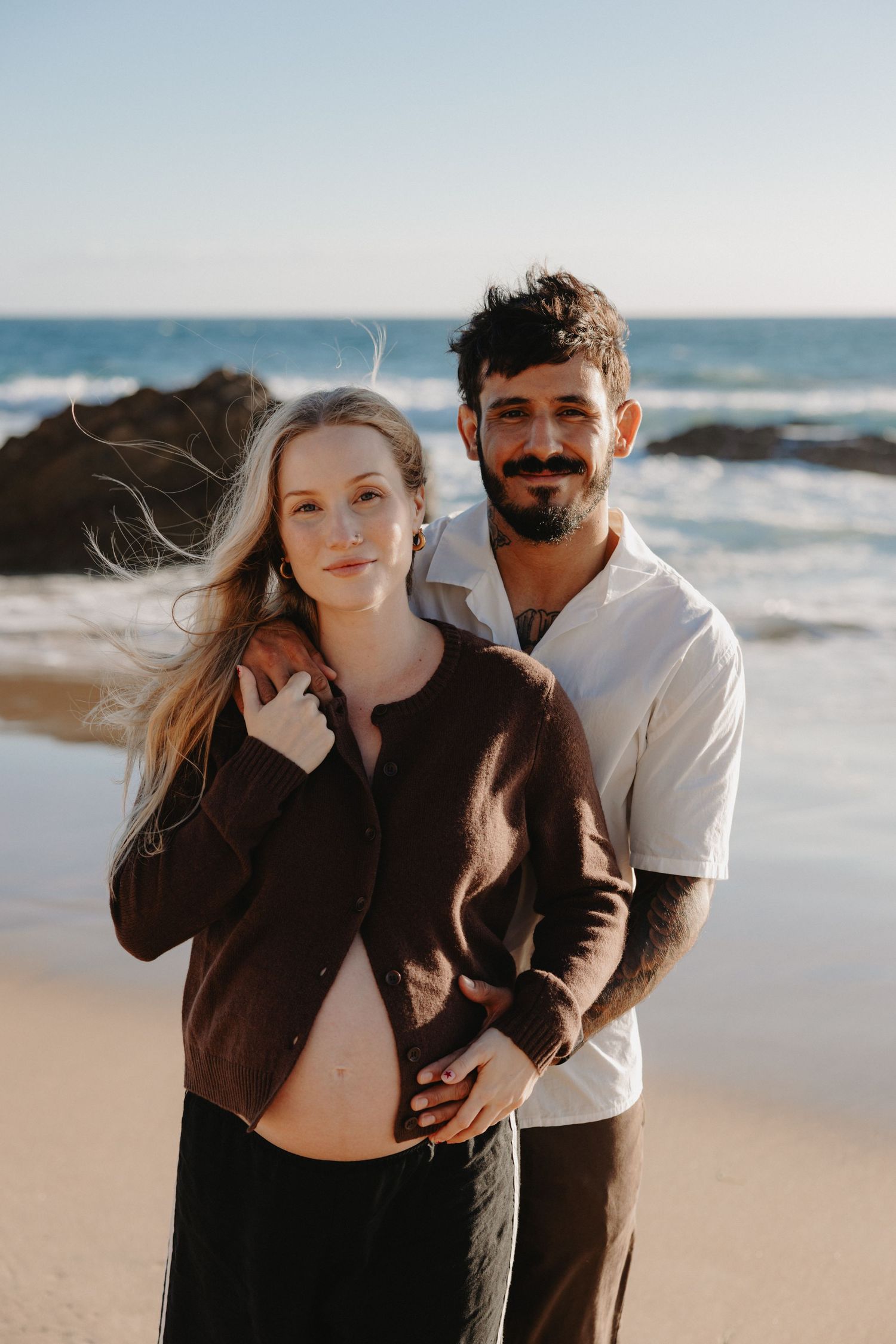 Romantic couple embraces on sandy beach at sunset with ocean waves and rocks in background.
