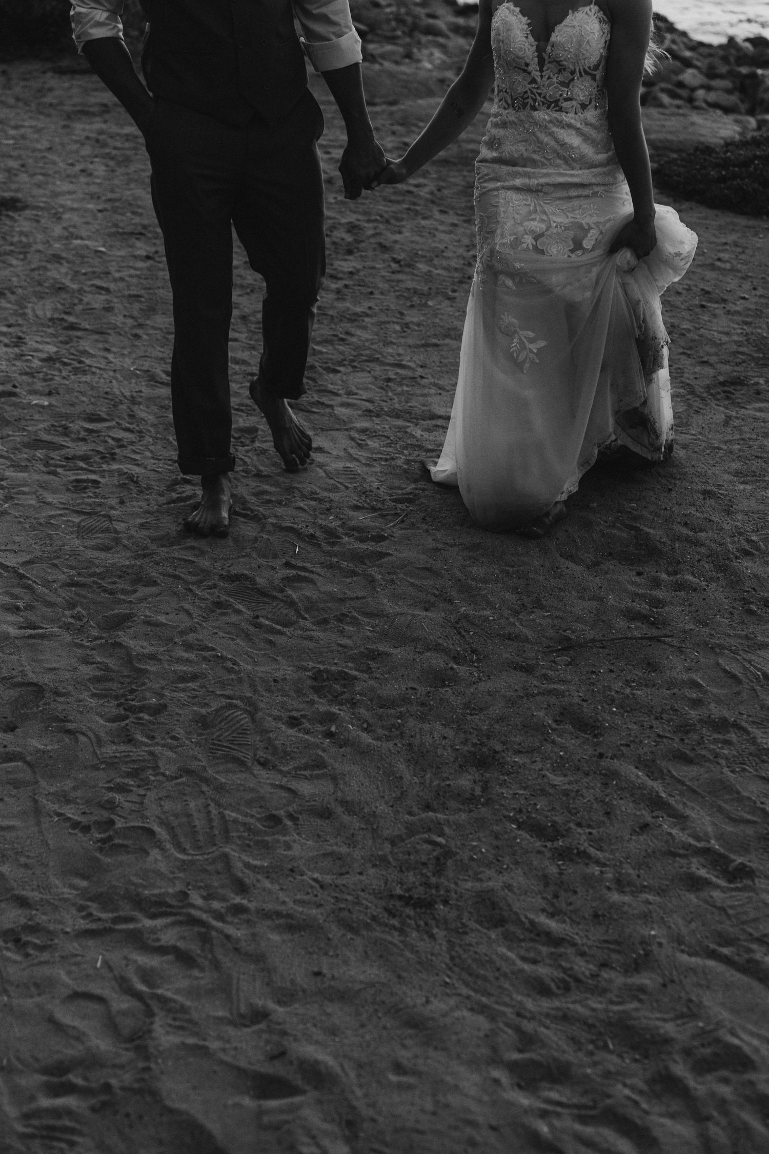 Black and white photograph showing people walking on a sandy beach with formal attire and long dresses.