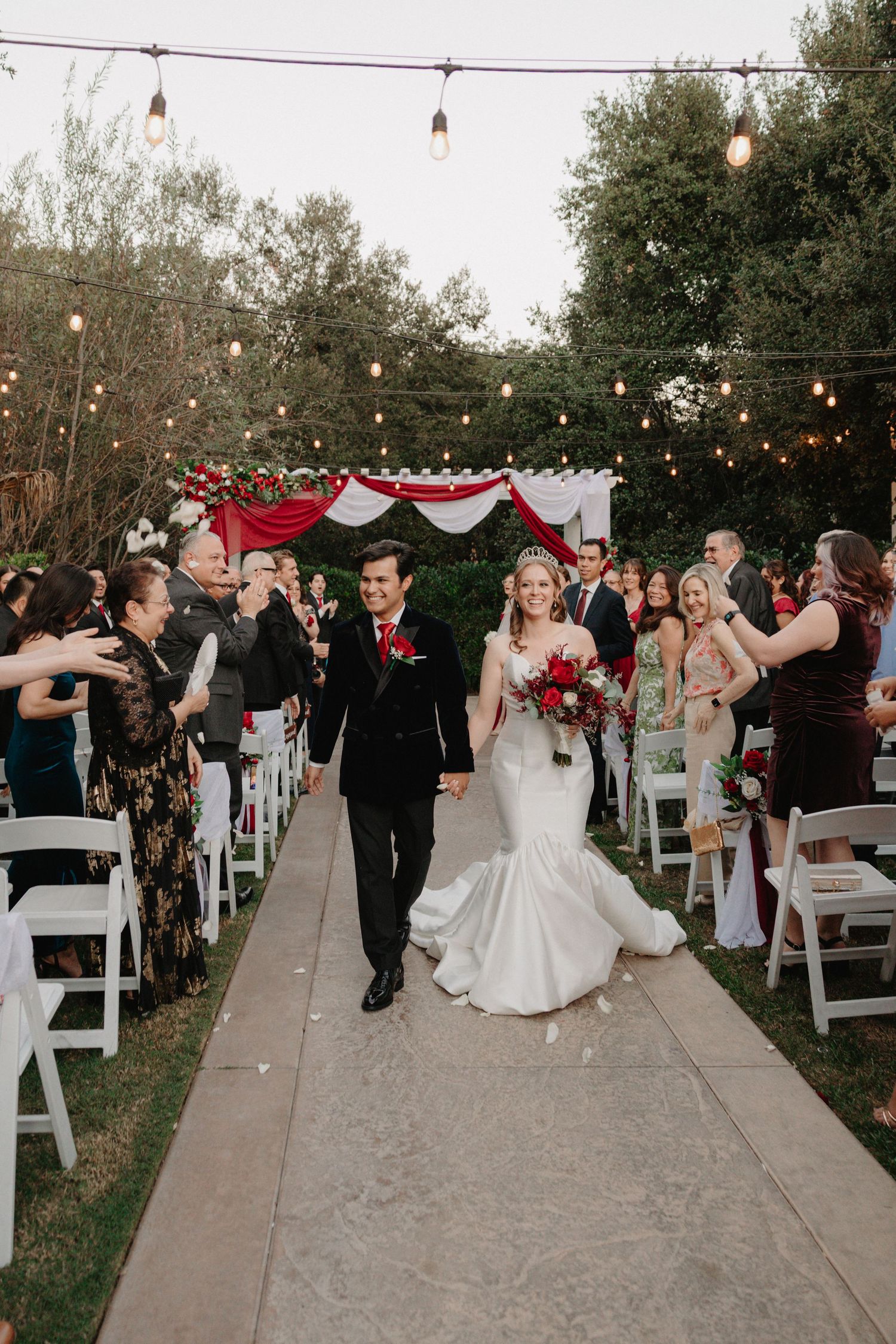 Wedding ceremony guests celebrate under string lights as newlyweds walk down outdoor aisle after exchanging vows.