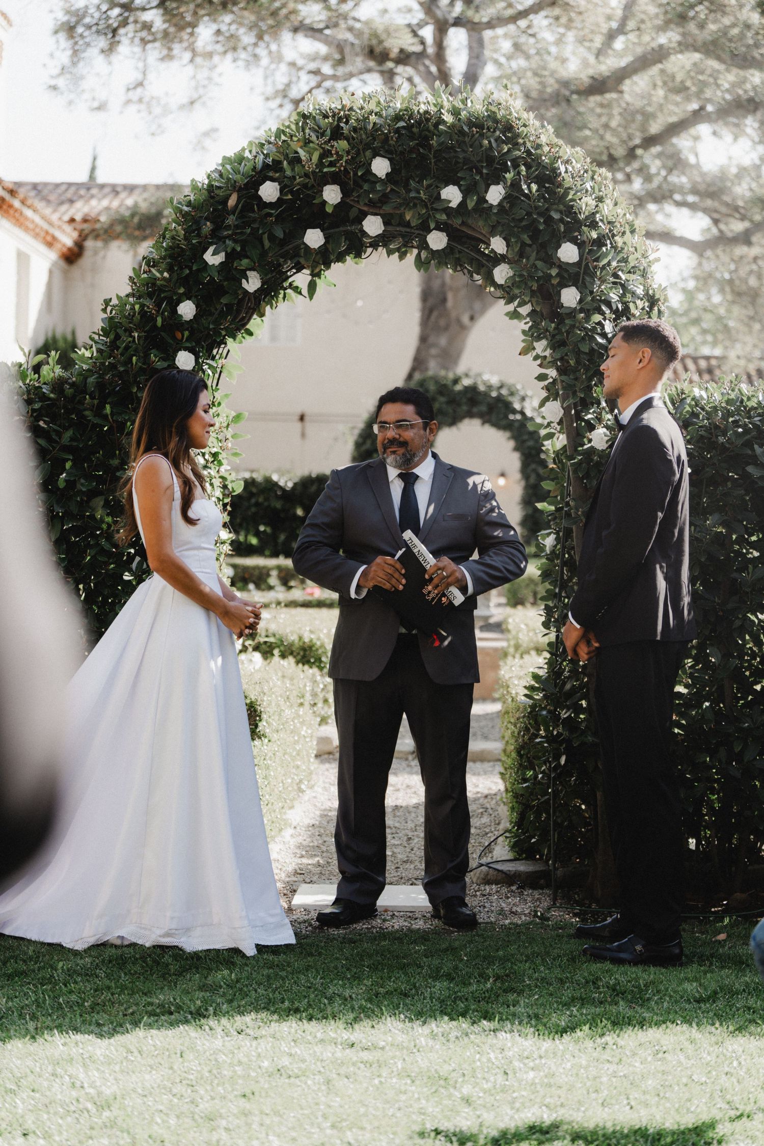 Wedding ceremony setup with floral arch and couple exchanging vows in an outdoor garden setting.