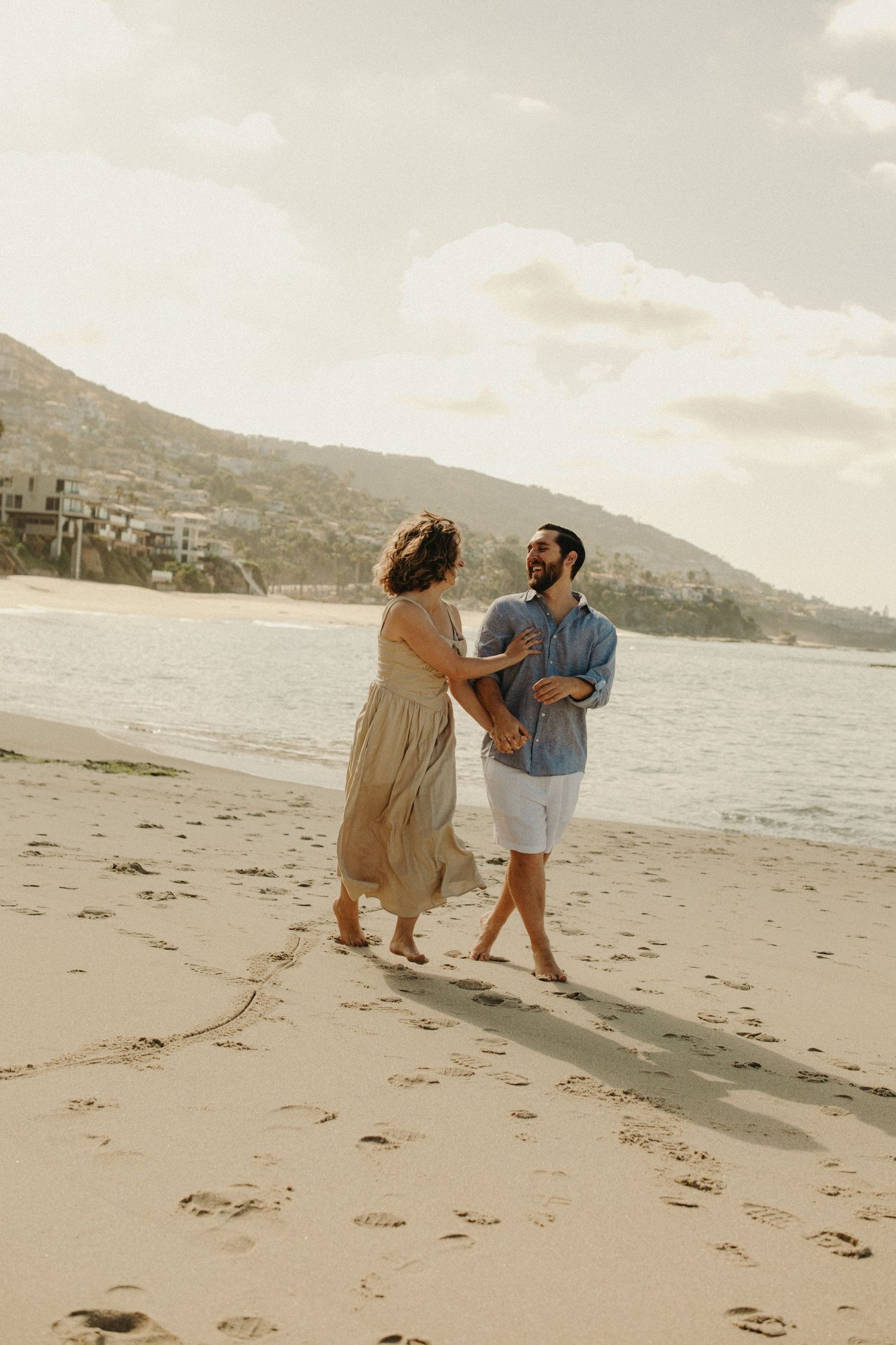Couple dancing barefoot on a sandy beach at golden hour with ocean and hills in the background.