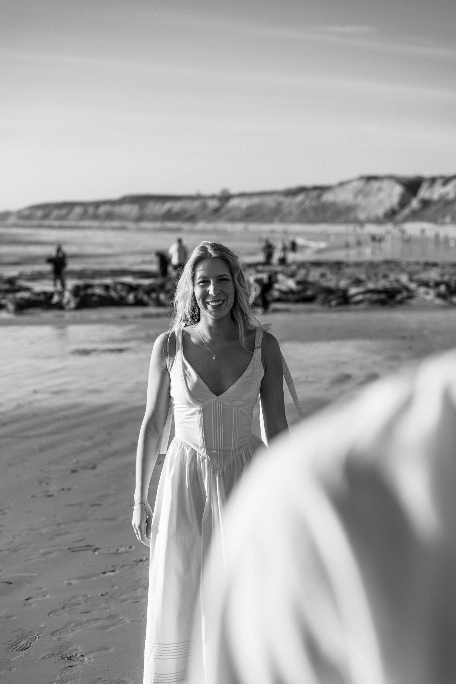 Person in flowing white dress walks joyfully along scenic beach with cliffs in background.