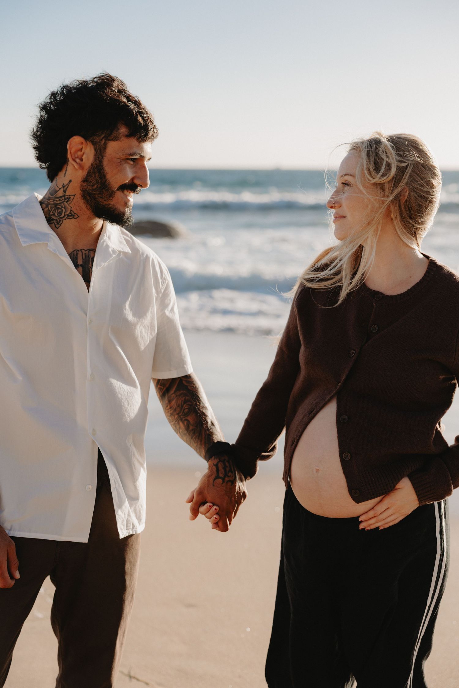 Romantic couple in black and white outfits walking together on a sandy beach with ocean waves in the background.