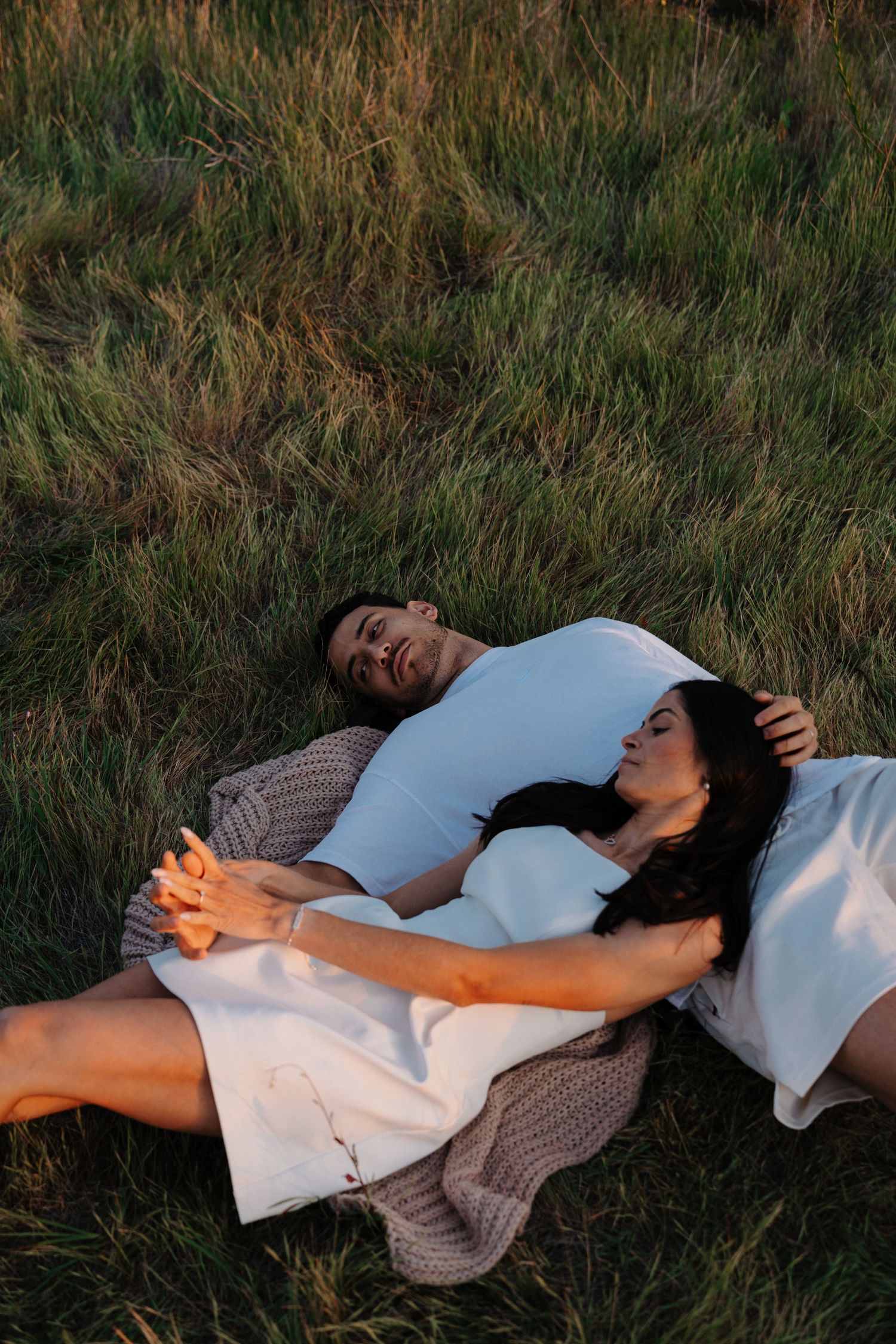 A couple relaxes together on a blanket in tall grass during golden hour, sharing a peaceful moment outdoors.