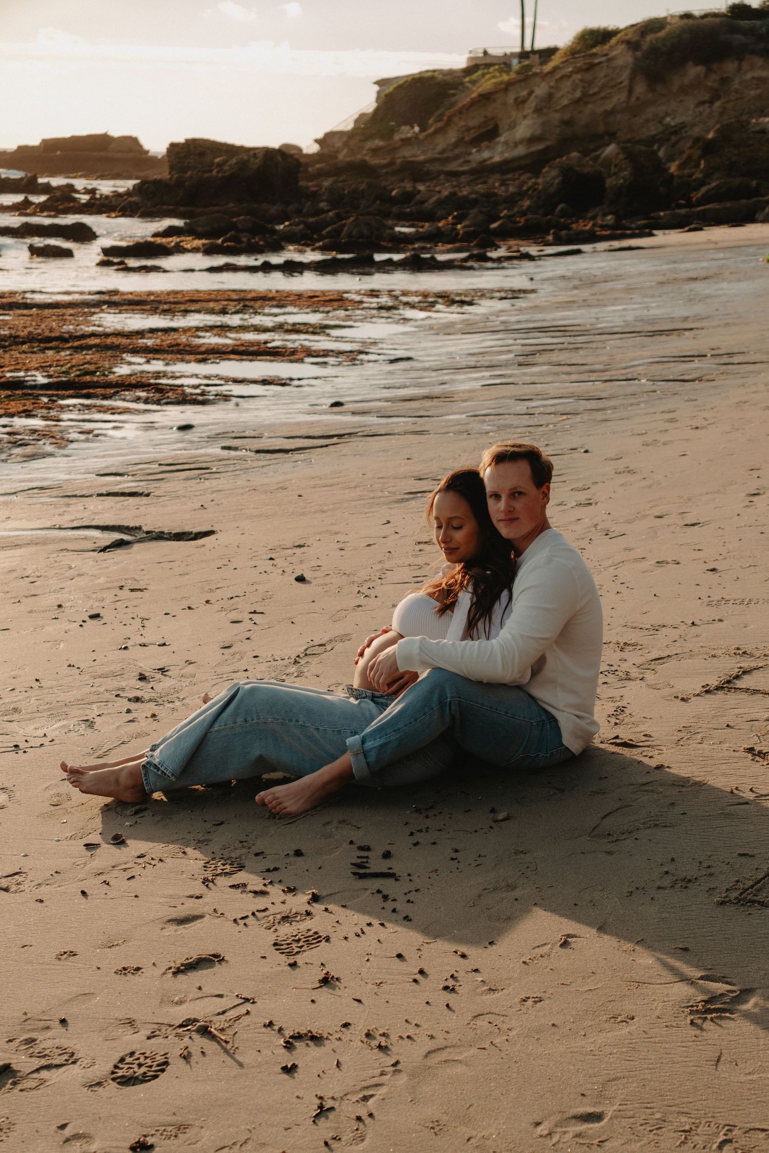 A couple embraces while lying on a sandy beach with rocky cliffs in the background during golden hour.