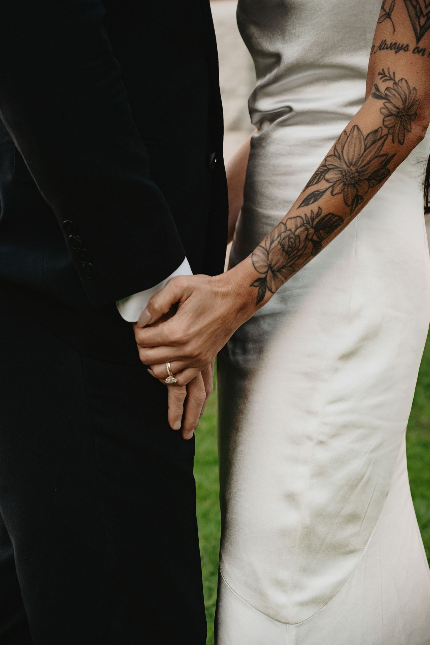 Close-up of couple holding hands showing floral tattoo sleeve against white wedding dress and black suit.