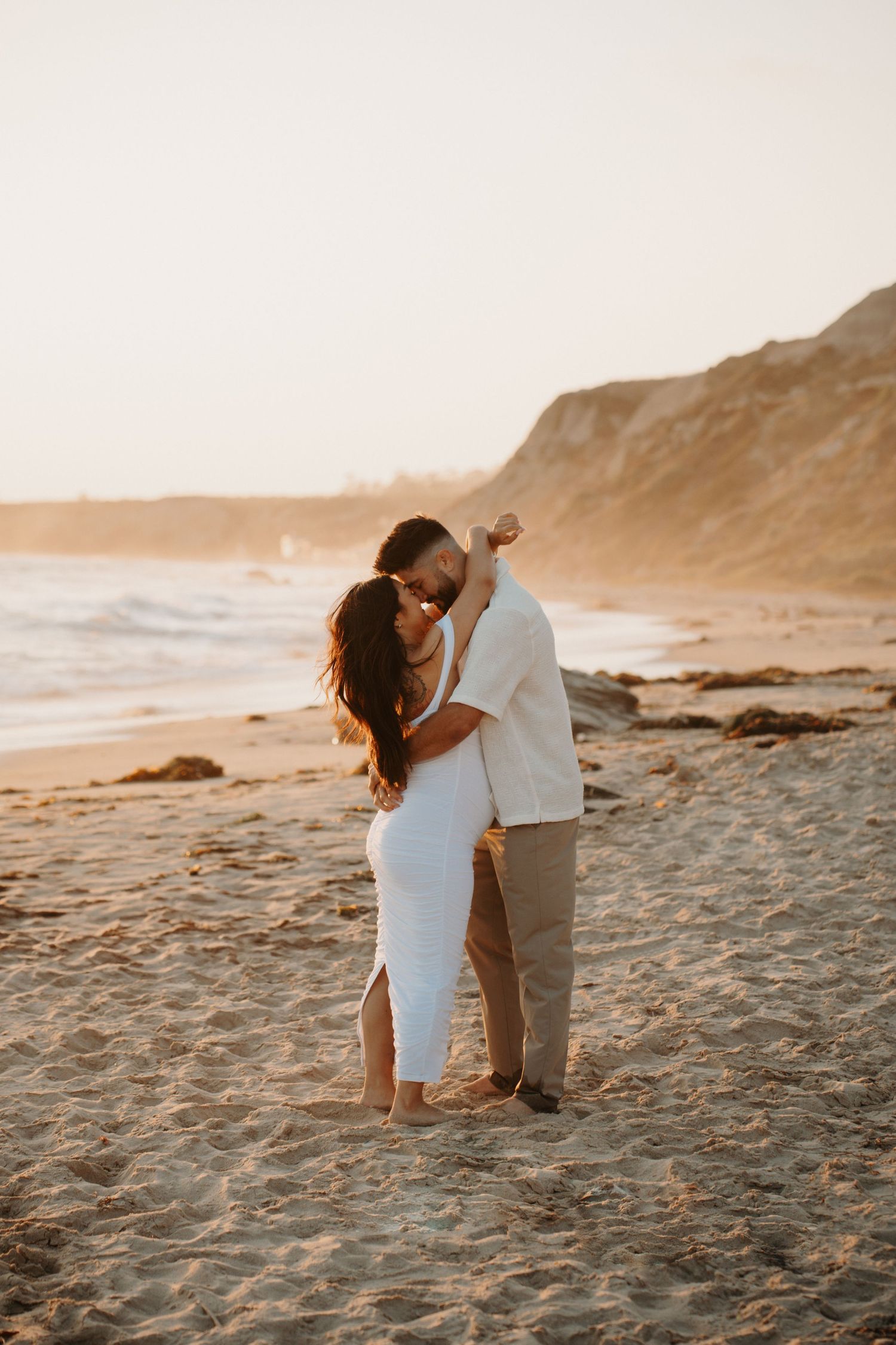 A couple shares a tender moment on a beach with cliffs in the background during golden hour.