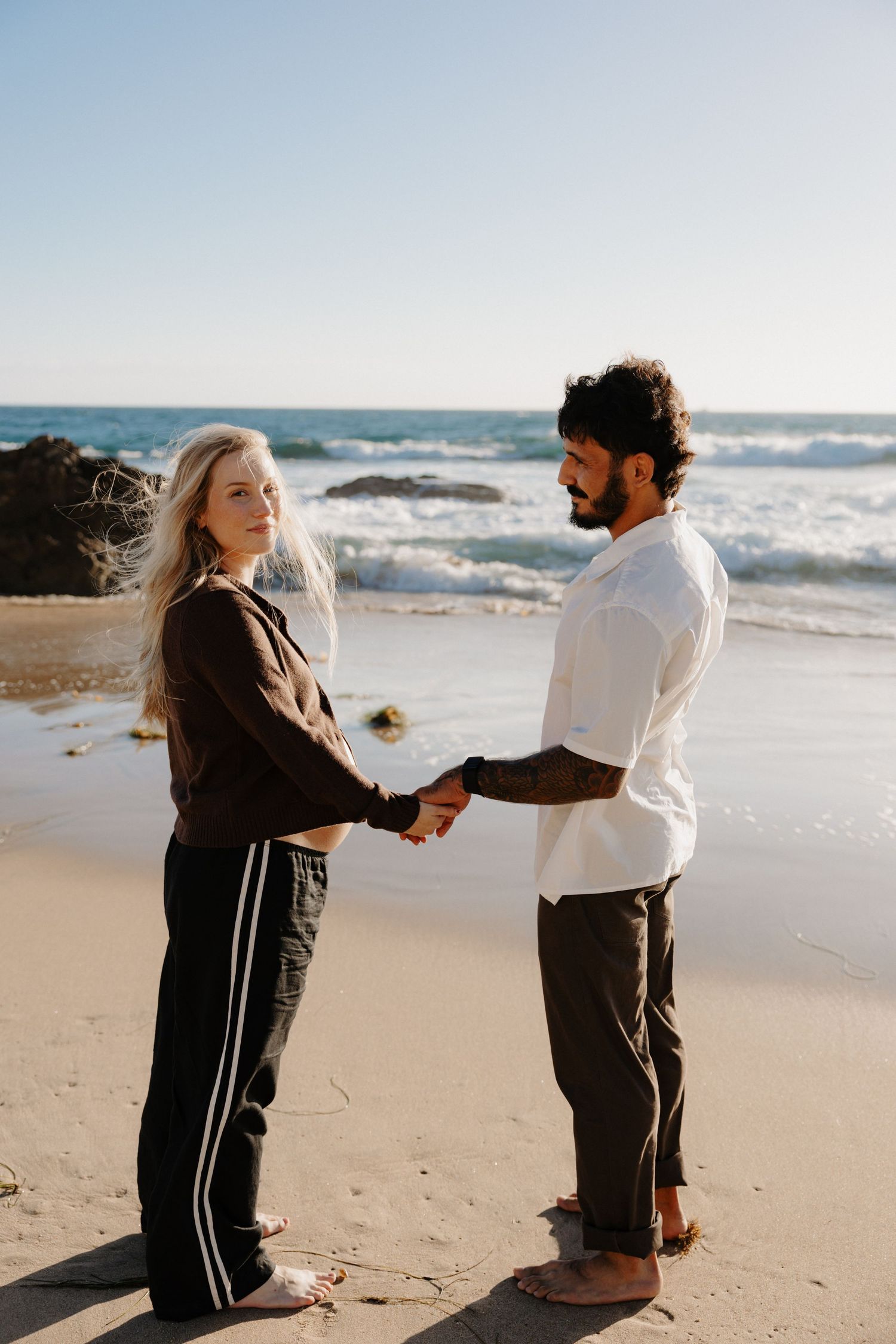 A couple holds hands while standing on a sunny beach with crashing waves and rocks in the background.