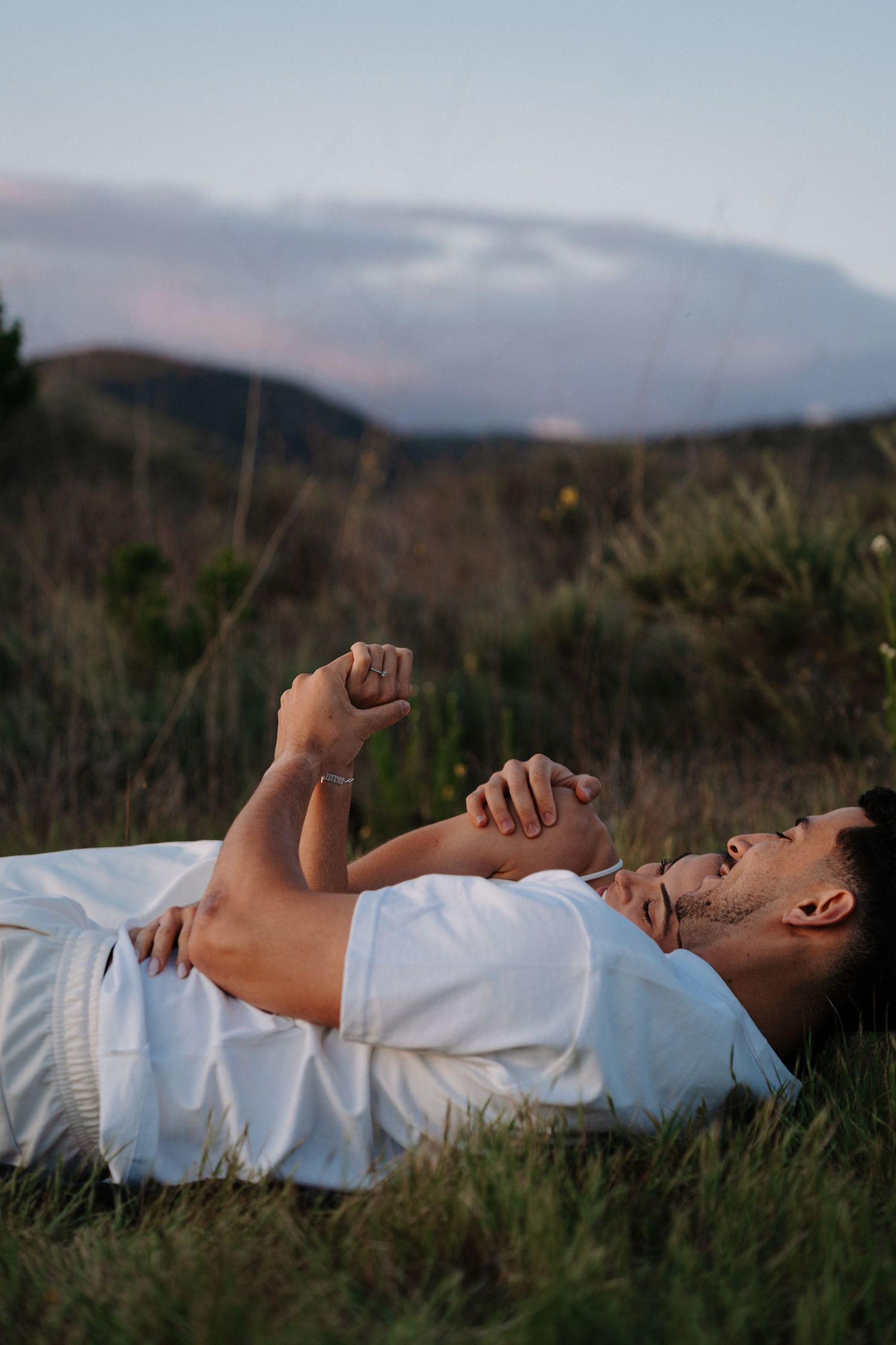 Person in white clothing lying peacefully in tall grass against a mountainous backdrop at dusk.