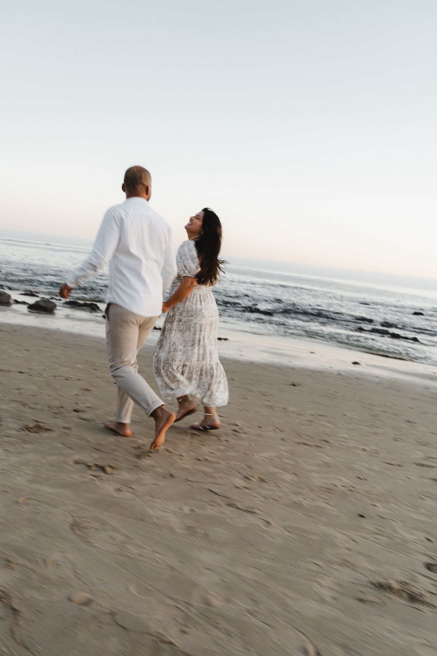 A couple in white outfits walks together along a sandy beach at sunset with ocean waves in the background.