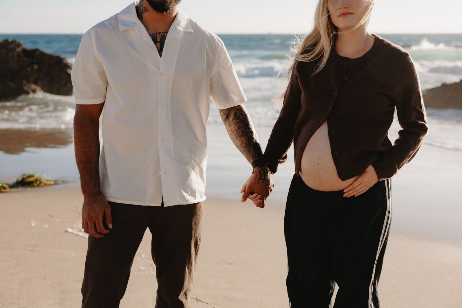 A couple walking hand in hand along a sandy beach wearing black and white outfits during a sunny day.