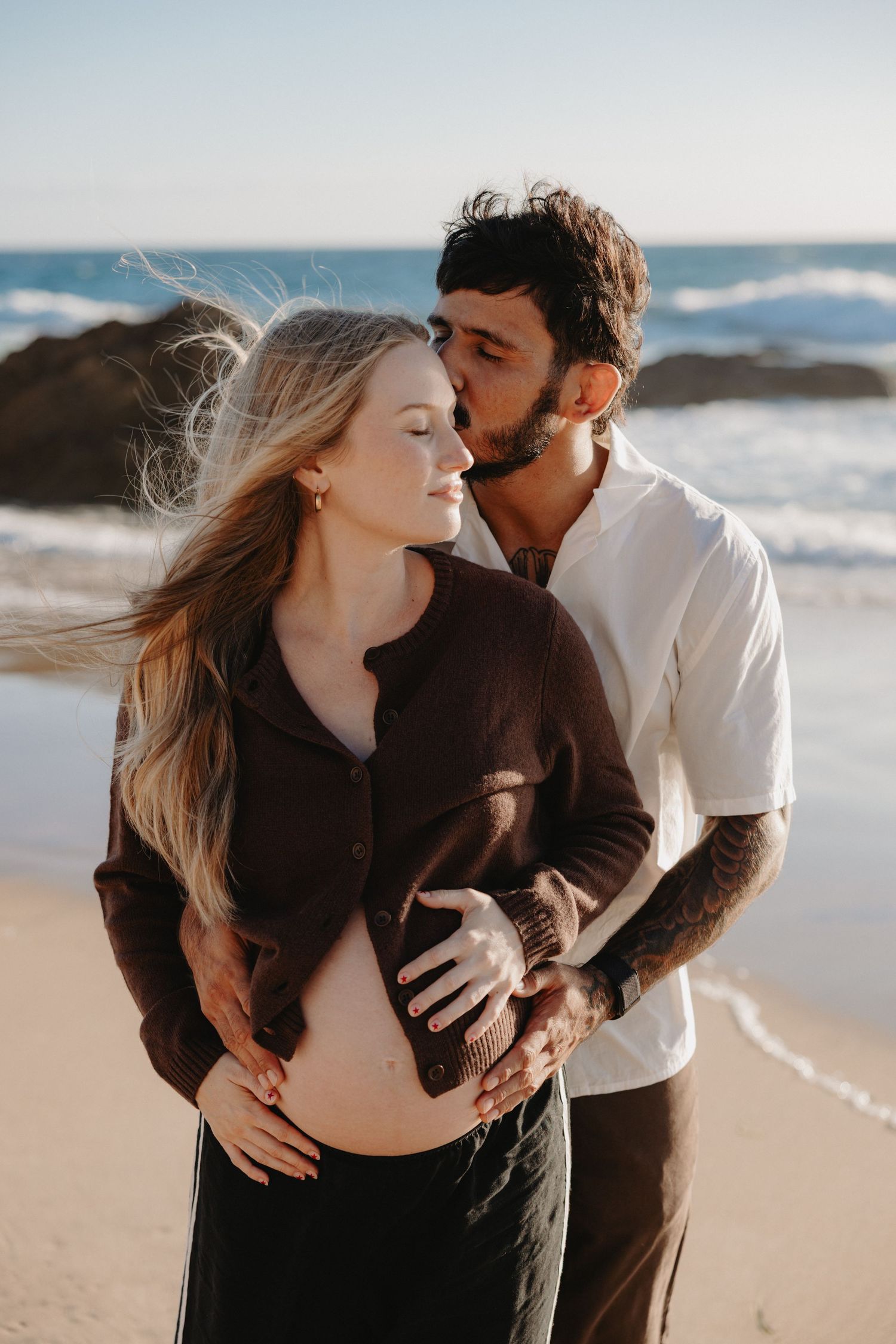 Romantic couple embraces on a sunny beach with waves crashing in the background.
