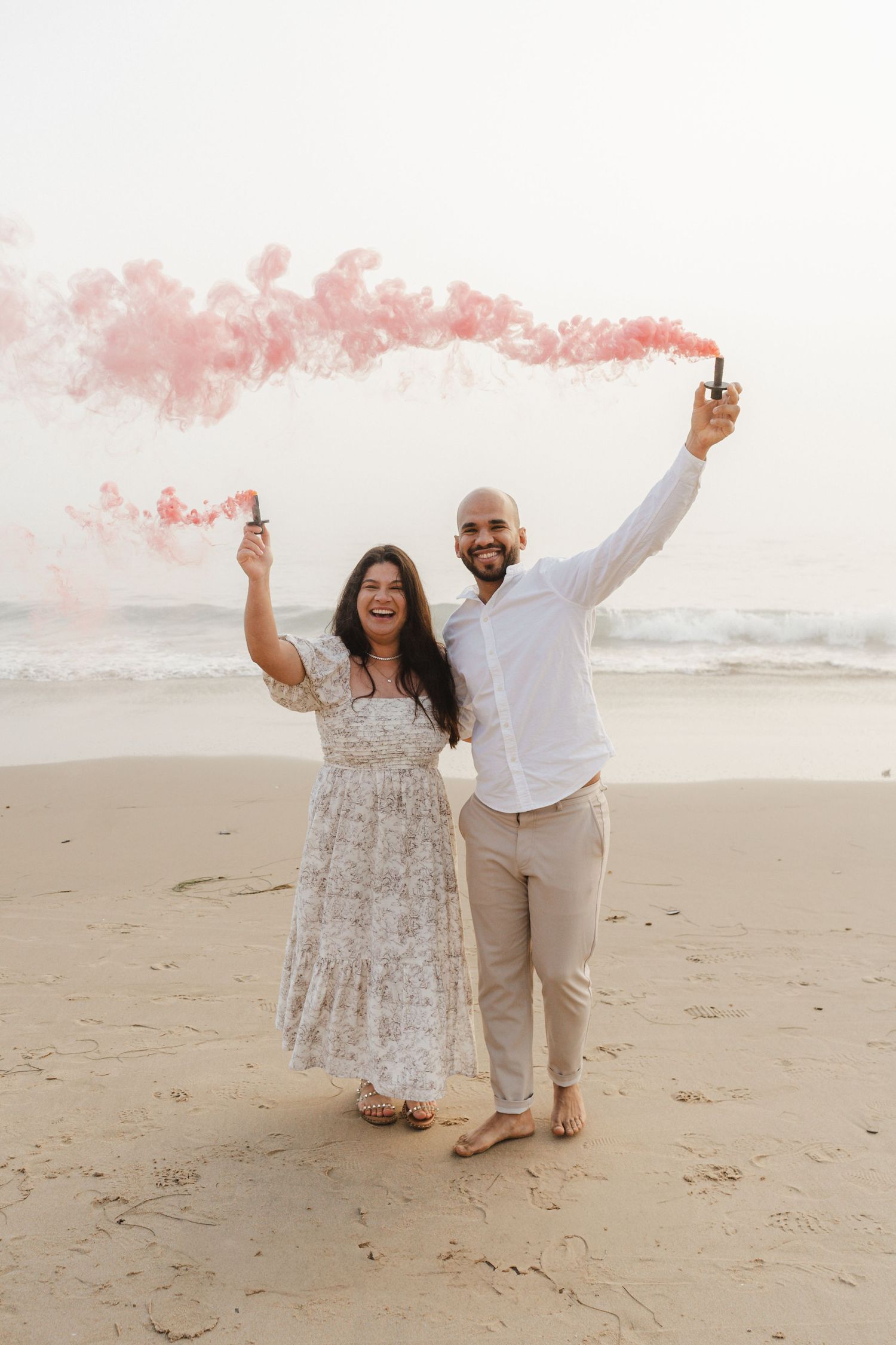 A couple celebrates on the beach holding pink smoke flares.