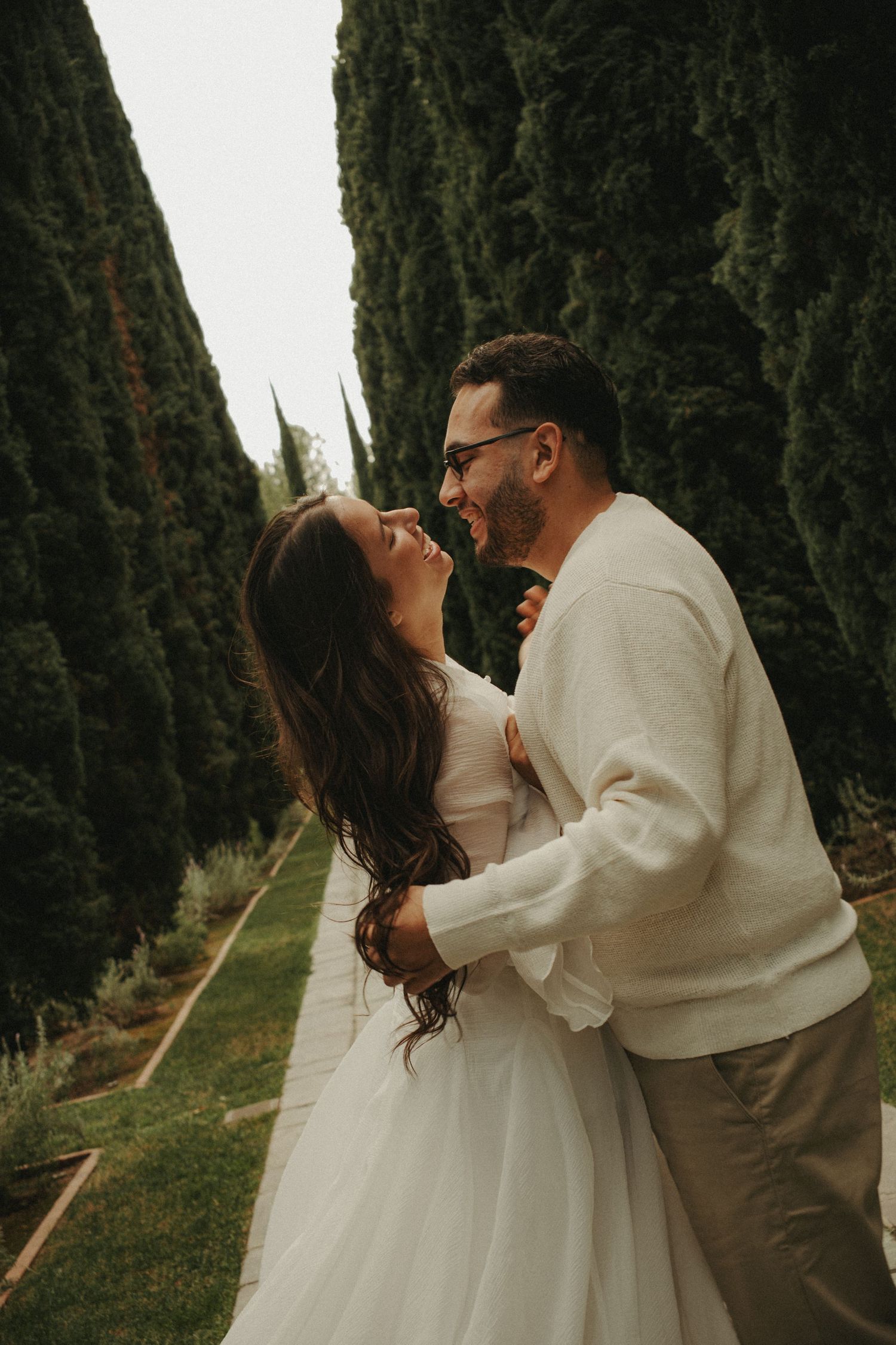 A romantic couple embraces in a garden pathway lined with tall cypress trees in warm evening light.