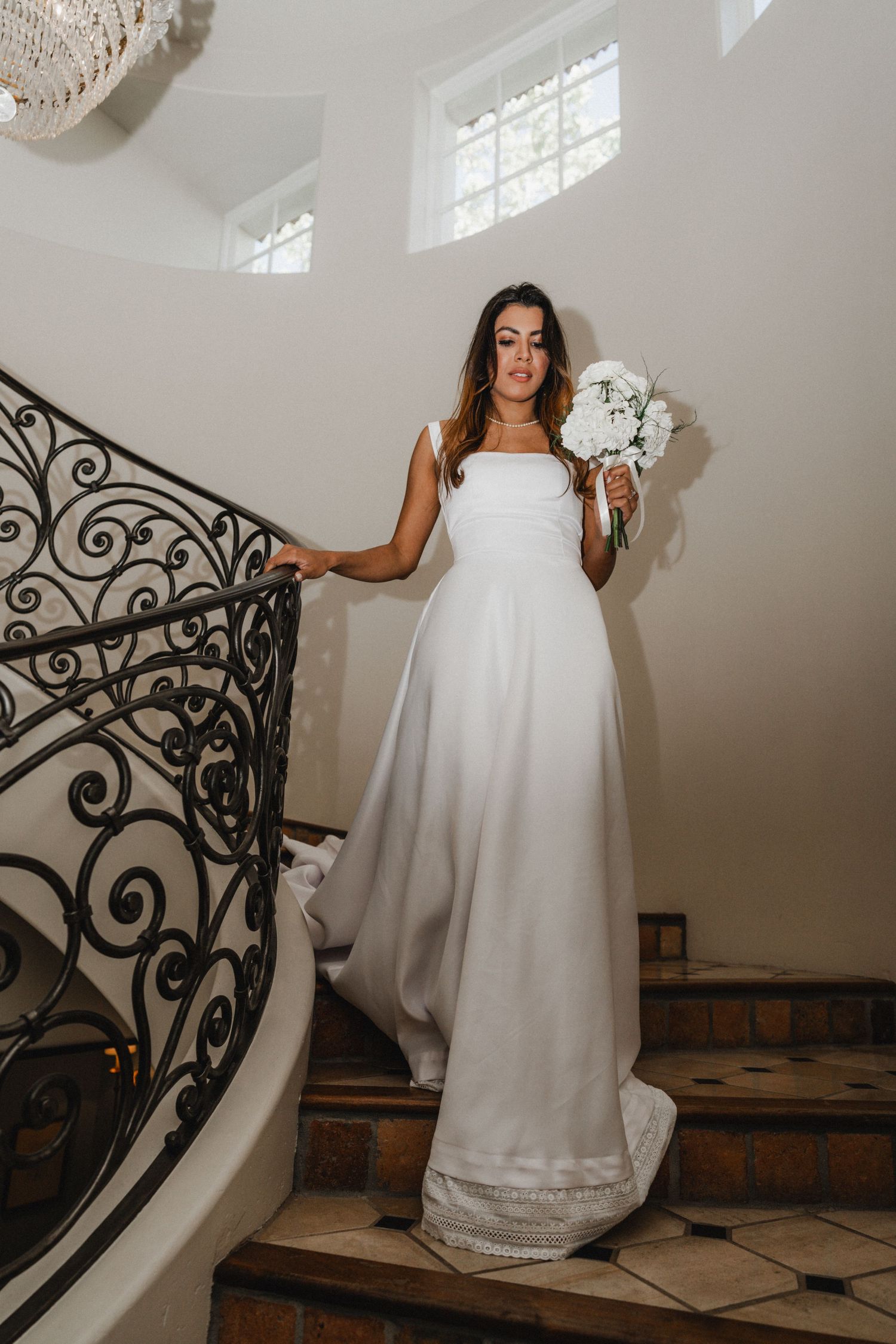 Person in flowing white wedding dress posing on elegant spiral staircase with ornate railing.